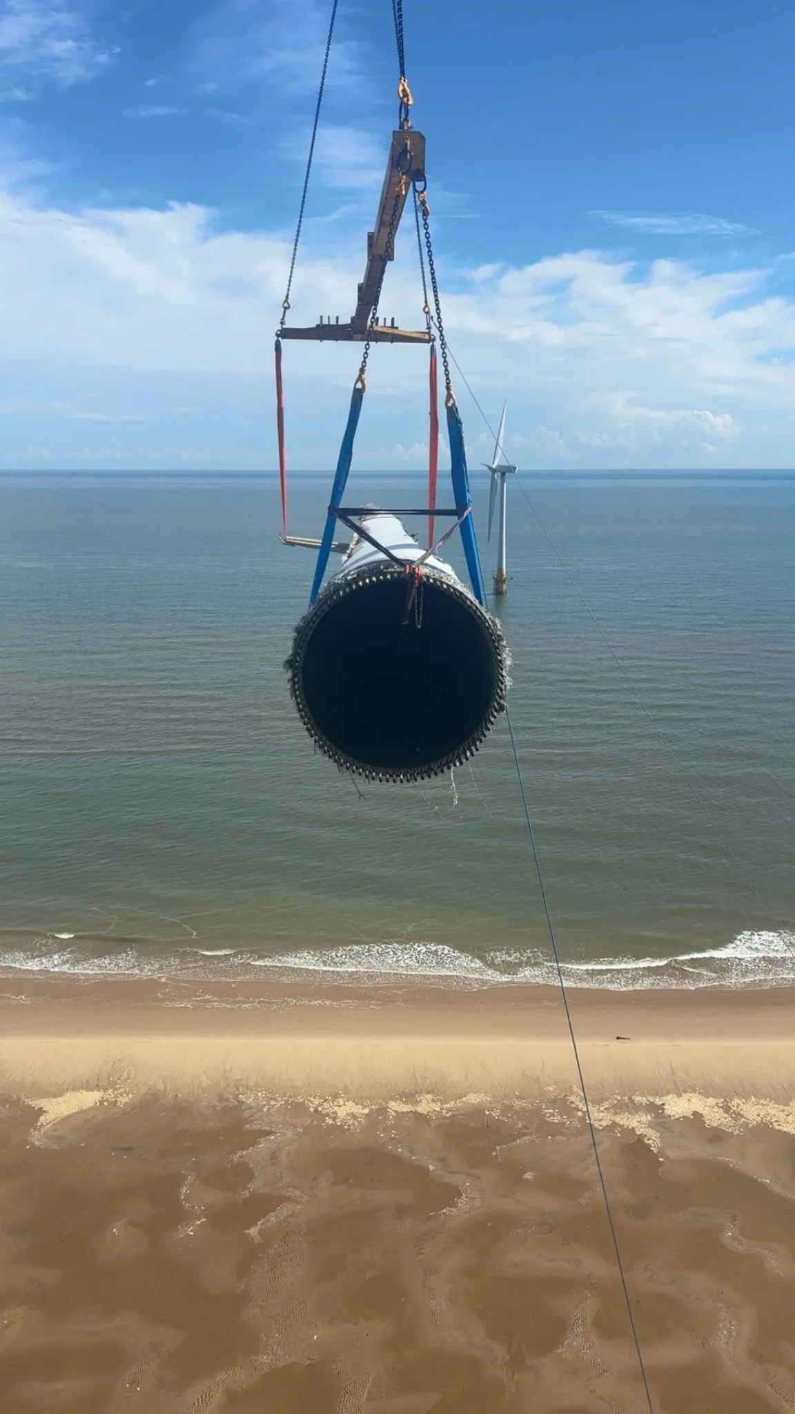 Horizontal view of a large wind turbine blade being installed over or near the beach, with the blue ocean and sky in the background.