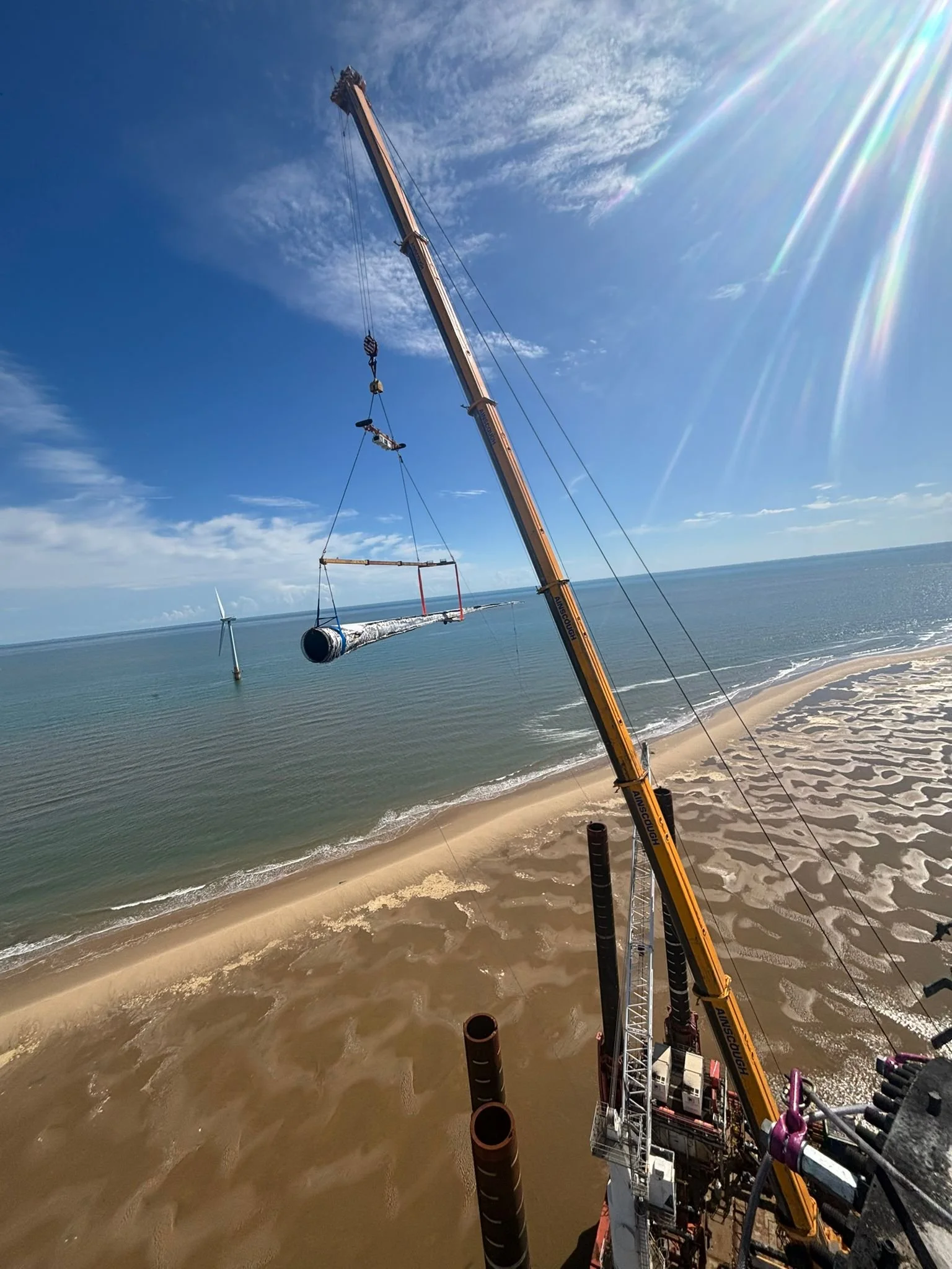 A construction crane lifting a large pipe on a beach with wind turbines in the water and a blue sky overhead.