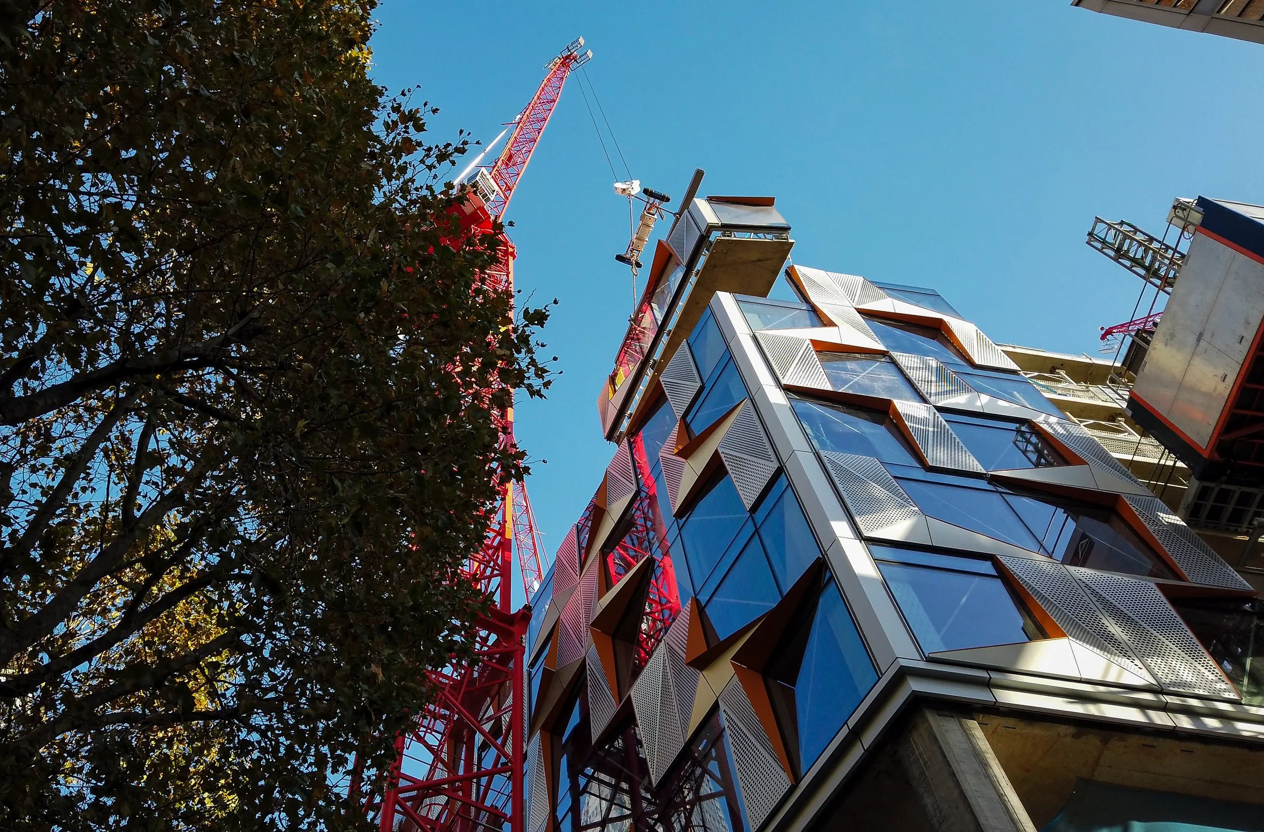 View of a modern building under construction with a red construction crane against a clear blue sky, and a tree on the left.
