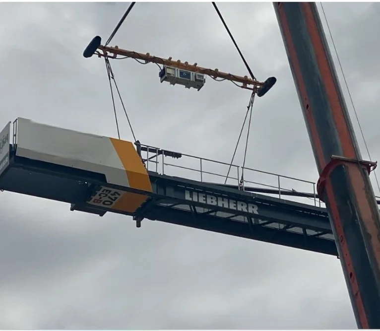 A construction crane lifting a piece of equipment or platform labeled "Liebherr." The crane's arm is extended upward against a cloudy sky.