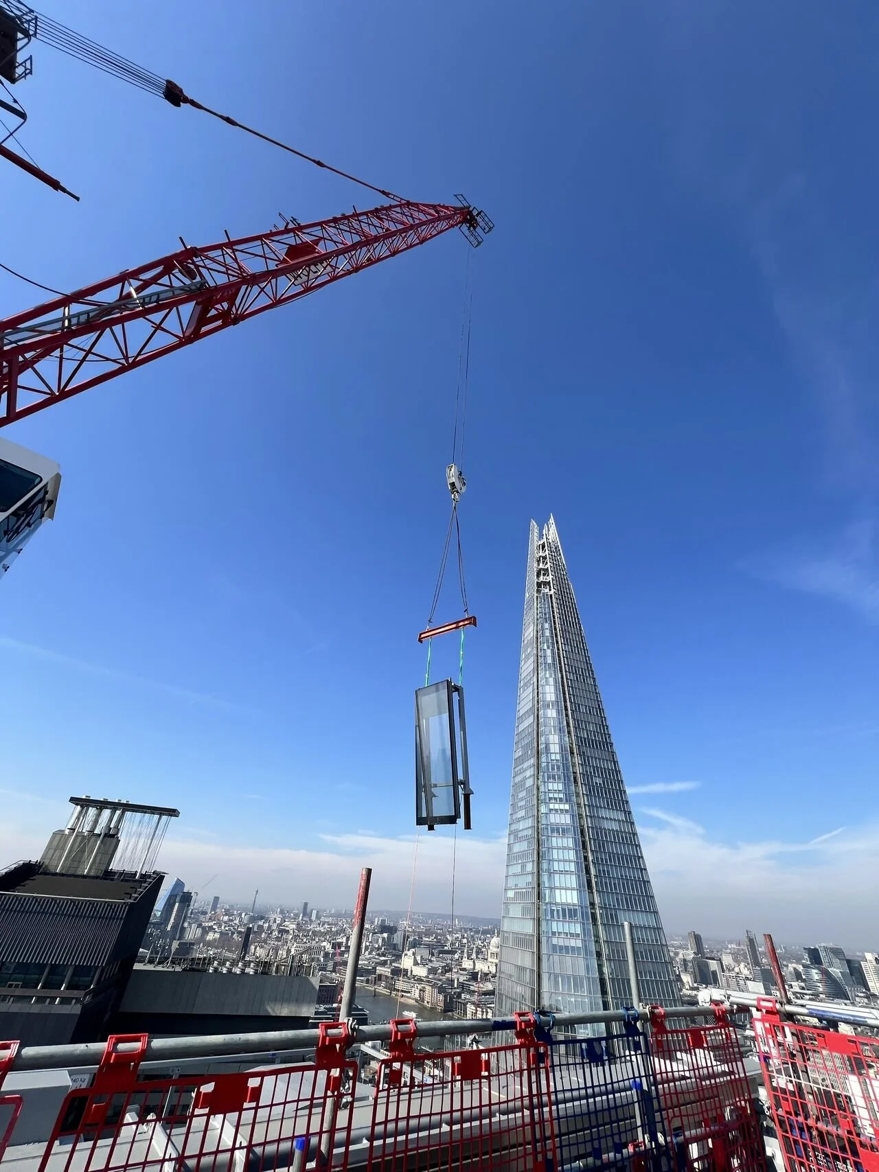 A construction site on a high-rise building in London with a crane lifting a glass panel, the Shard in the background, and a city skyline under a clear blue sky.