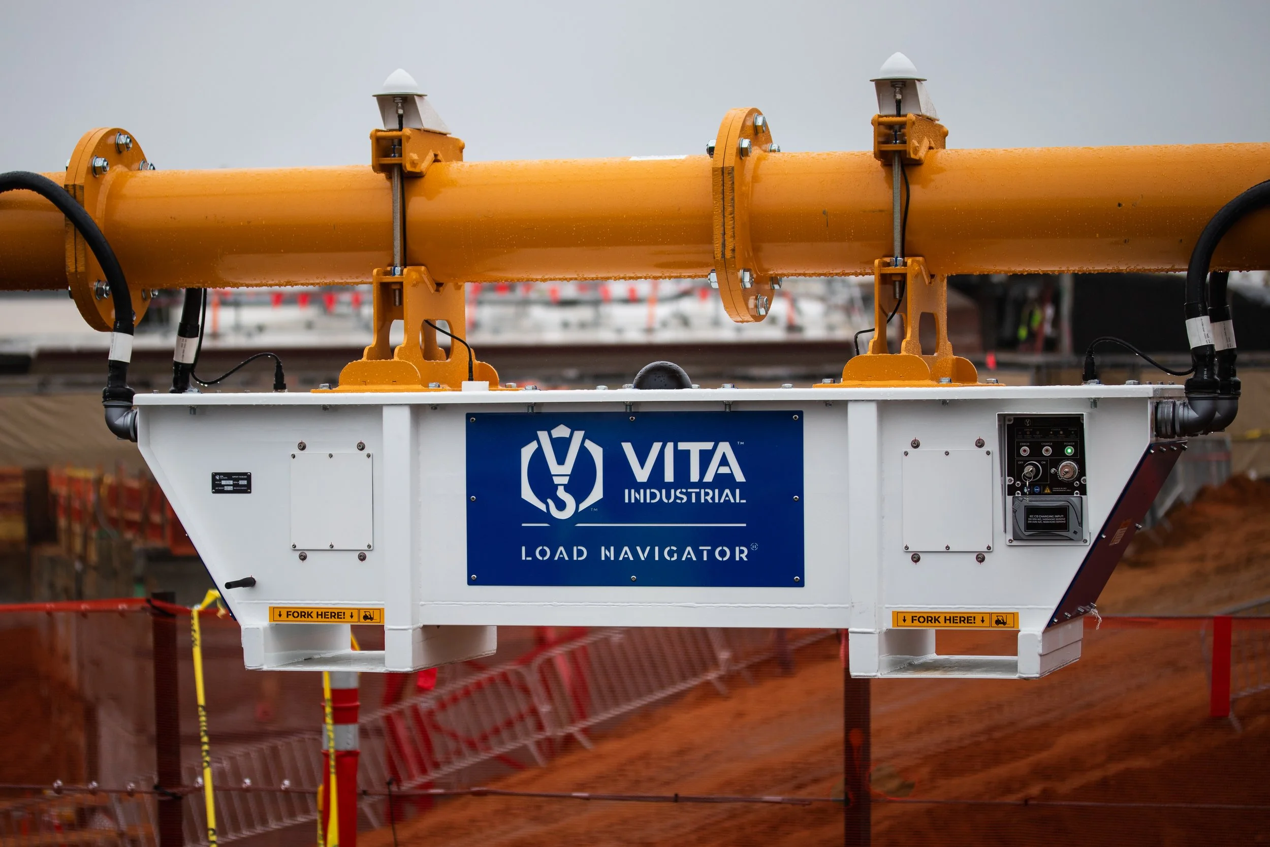 A construction site overhead load hosting equipment labeled 'Vita Industrial Load Navigator,' with a large orange pipe attached, surrounded by construction barriers and equipment.