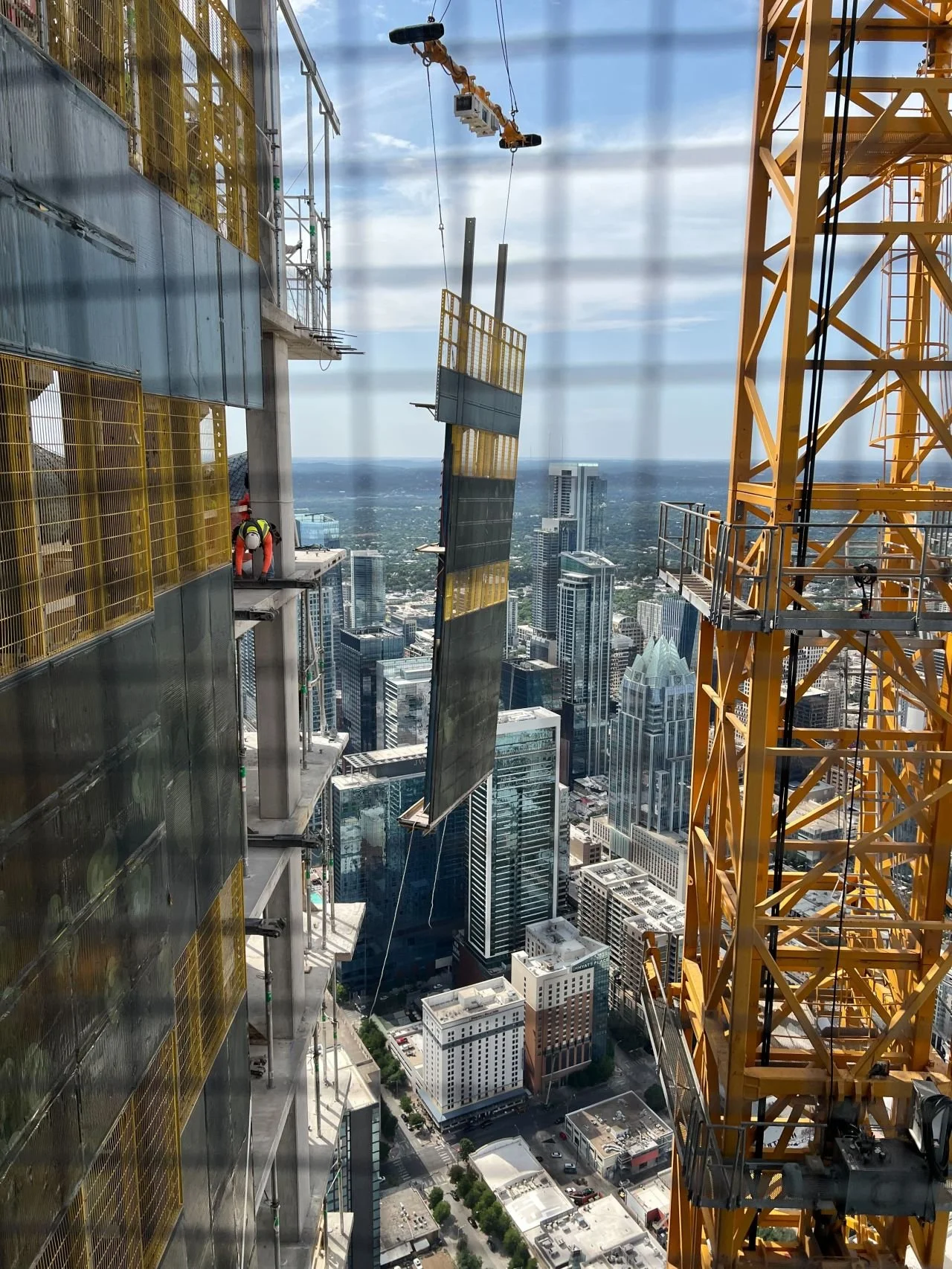 A construction worker on a building under construction high above a city skyline, with a crane to the right and a cityscape with tall buildings and streets below.