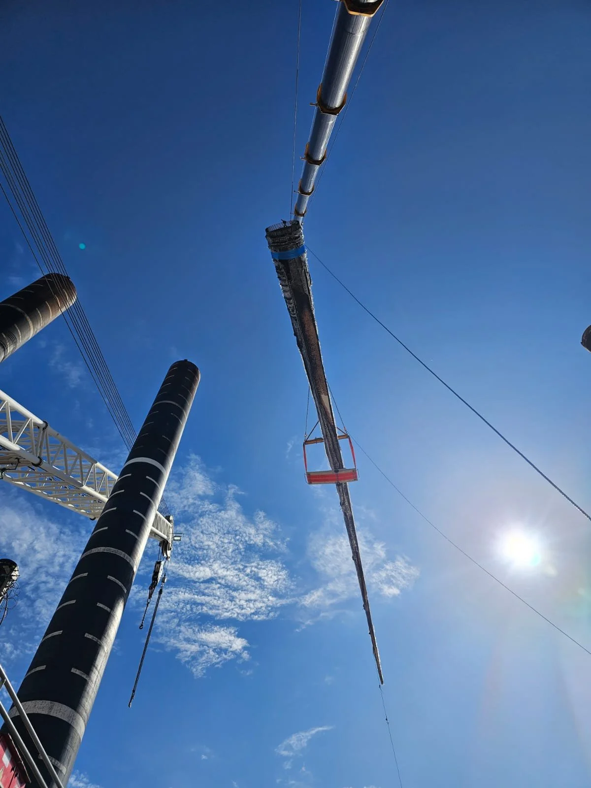 A crane lifting a long, black, and red construction beam high into the blue sky with some white clouds and the sun shining brightly.