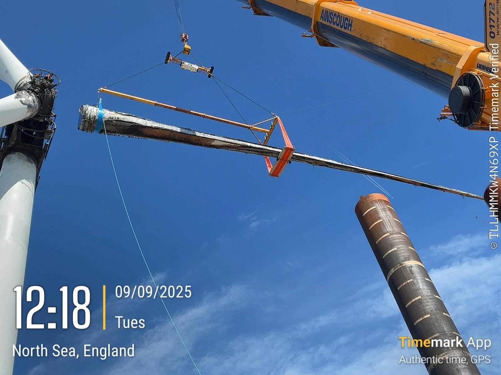 Workers are lifting a large section of a wind turbine blade using a crane. The blades are part of a wind turbine being assembled in North Sea, England, on a clear day.