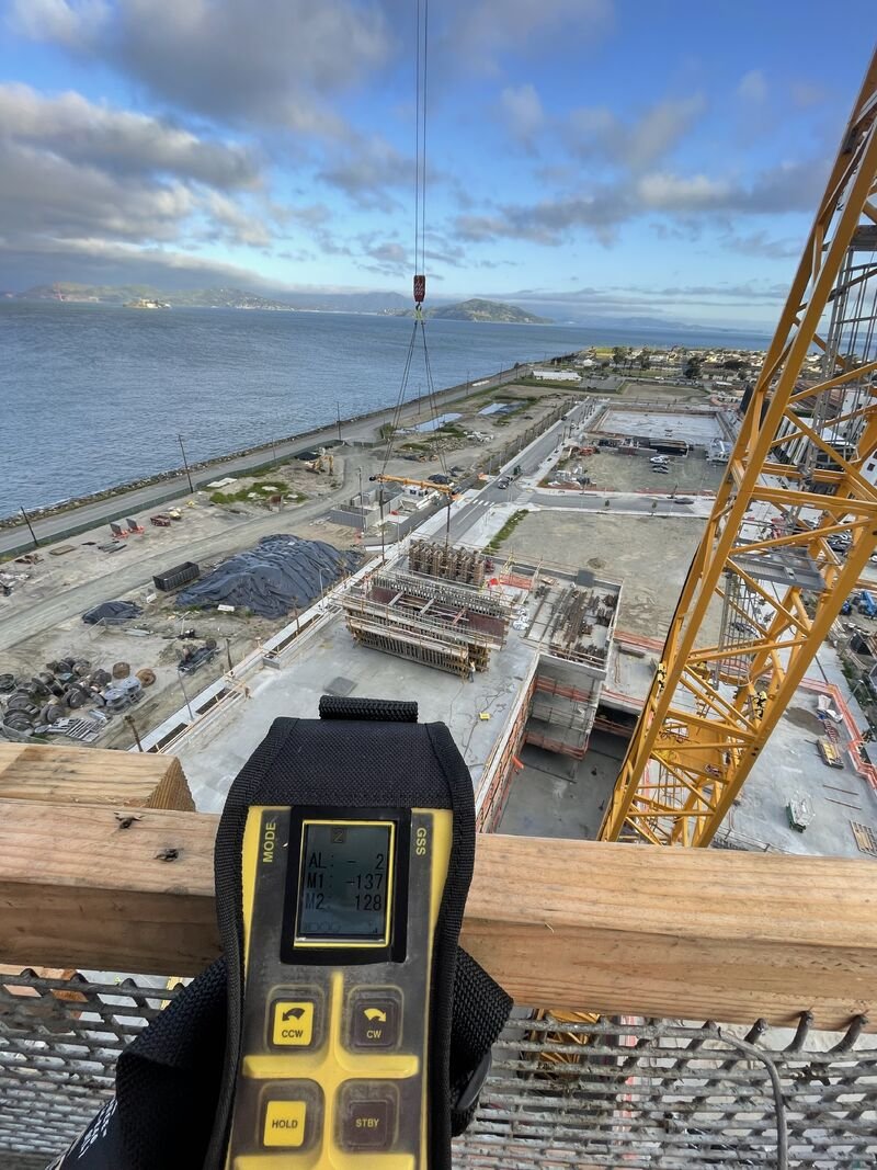 View from a construction site tower overlooking a body of water with a distant landmass. A yellow crane is visible, along with building structures and construction materials. Foreground shows a person holding a digital device with a measurement readi