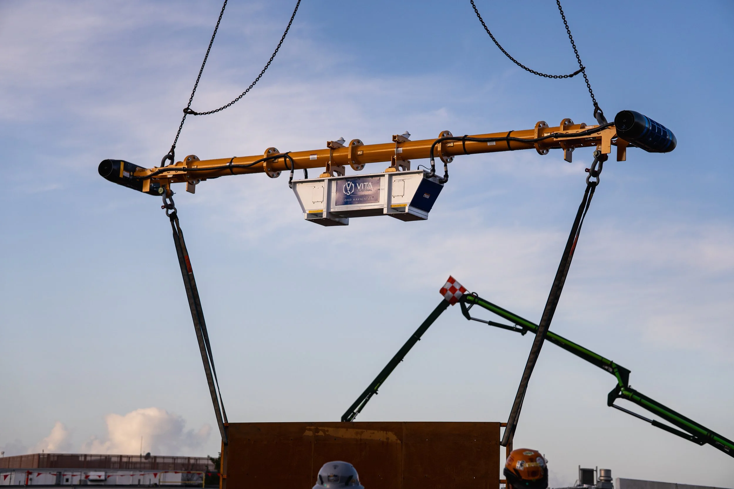 Construction or industrial equipment suspended in the air, including a yellow crane-like structure with chains, a white box with logo, and a green extendable arm, against a blue sky with some clouds.