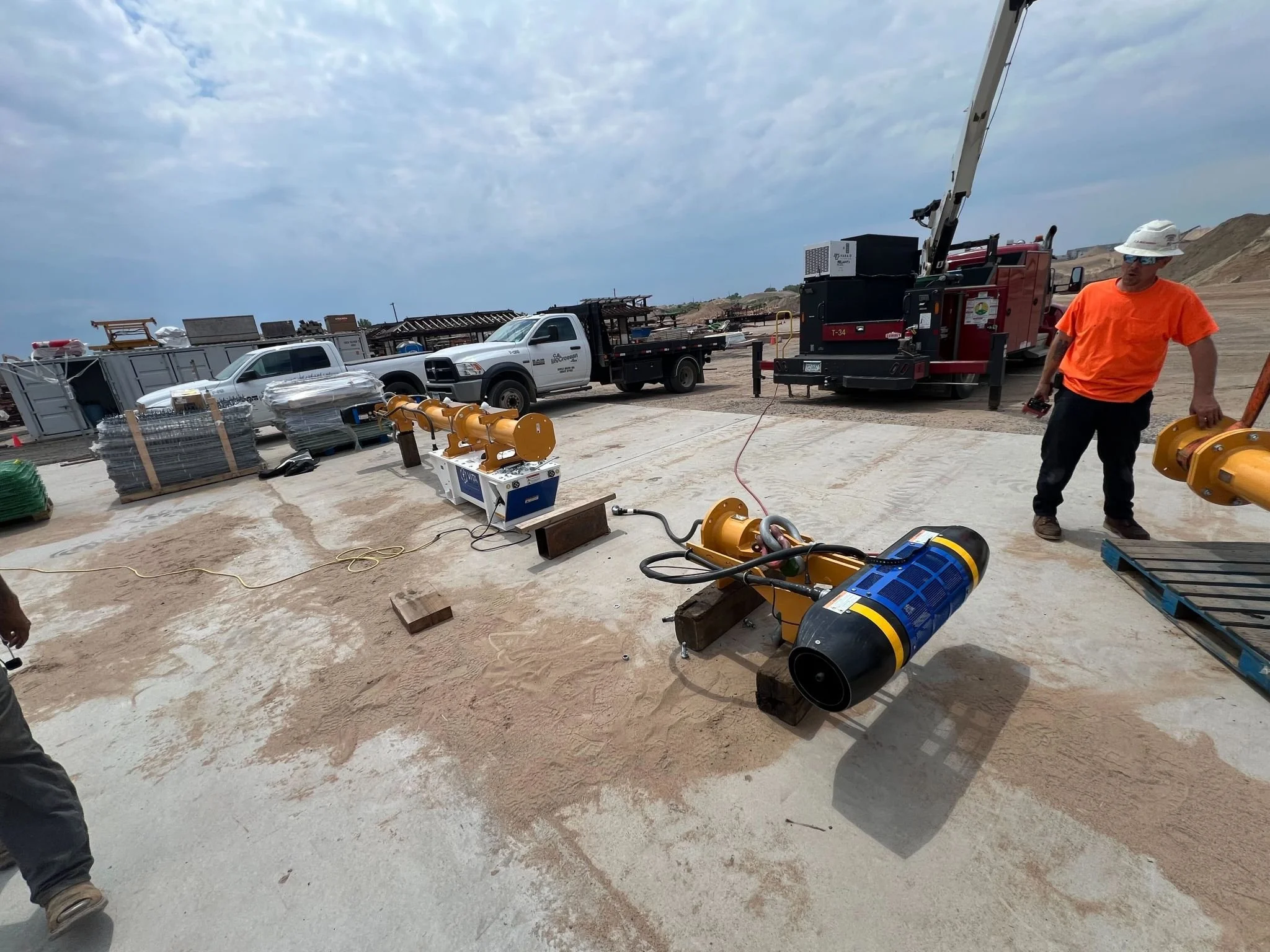 Construction site with machinery, trucks, and a worker in an orange shirt and white hard hat.