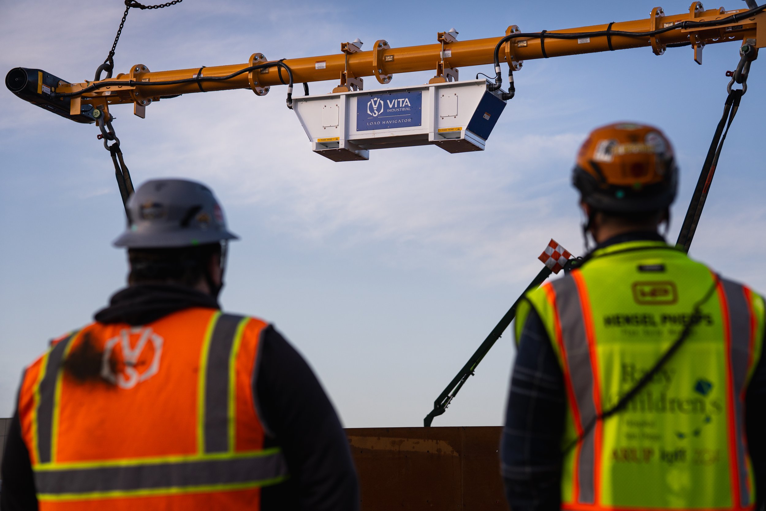 Two construction workers in safety vests and helmets look at a large orange crane with a white load holder marked 'VITA Industrial Load Navigator' against a cloudy sky.