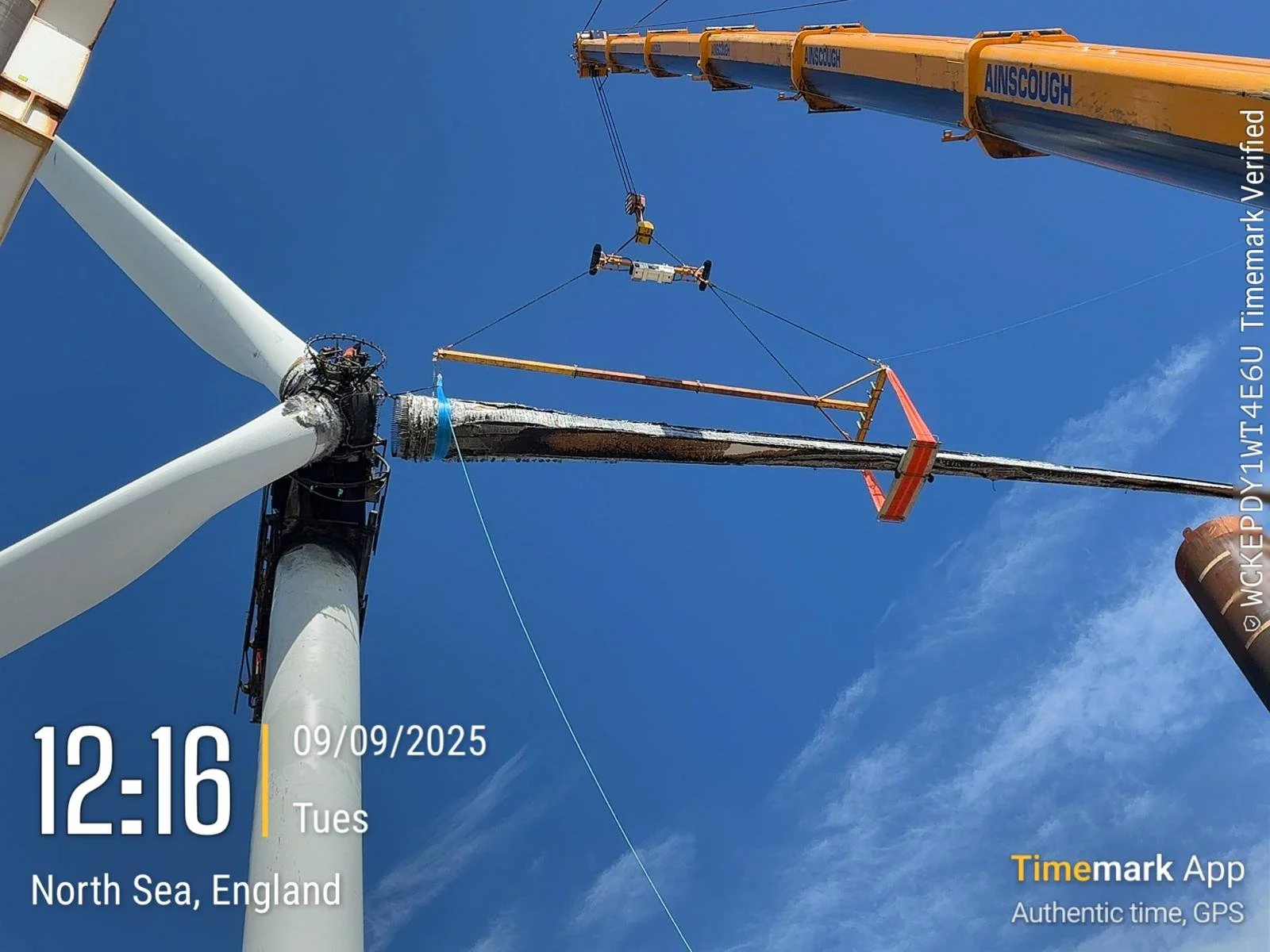 A wind turbine under maintenance with a crane lifting equipment to the turbine's top during daytime. Clear blue sky in the background.