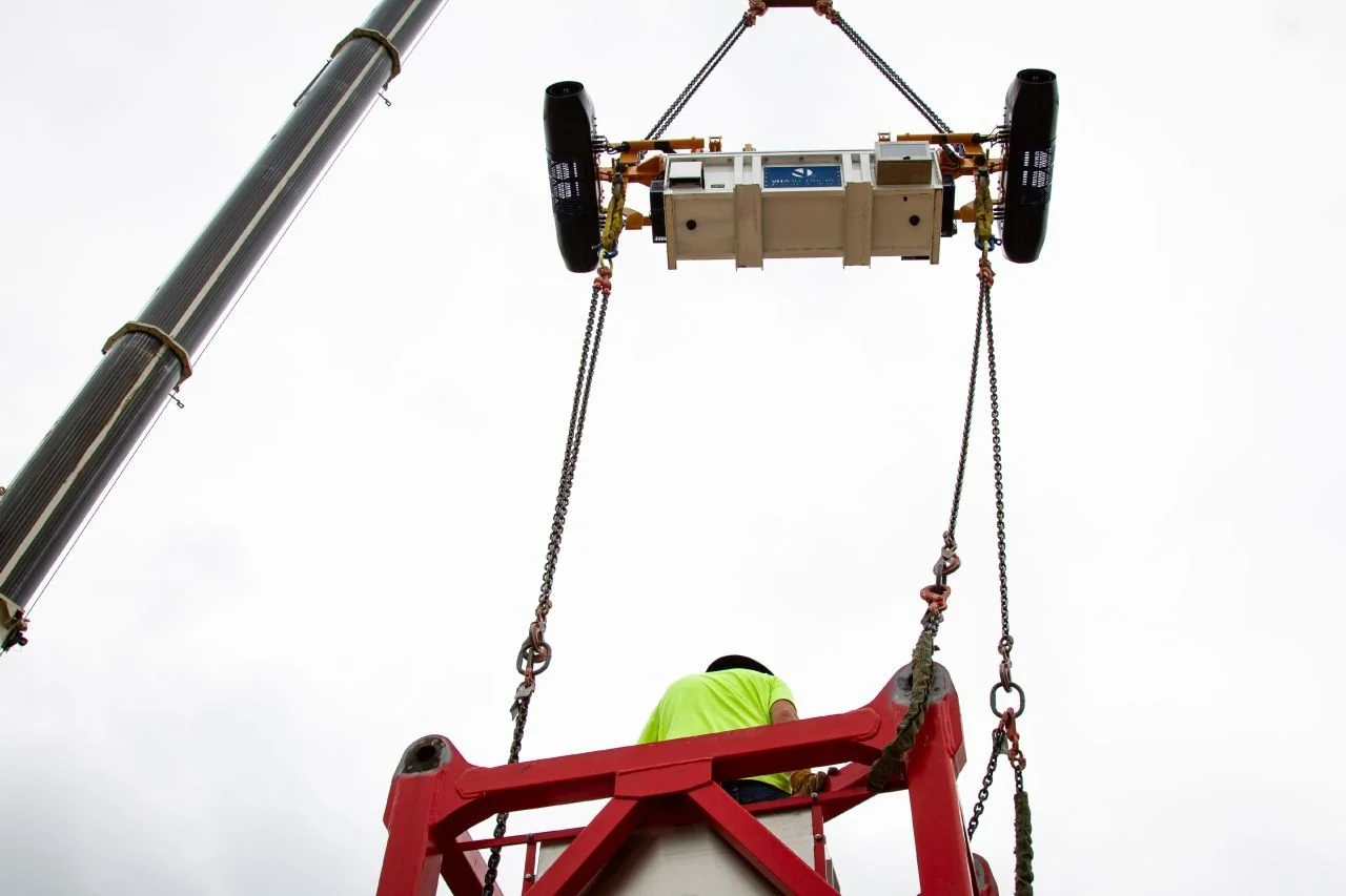 A worker in a yellow safety vest is on a scissor lift, installing or maintaining a large, square device using a crane with a long arm. The background is overcast, with a gray sky.