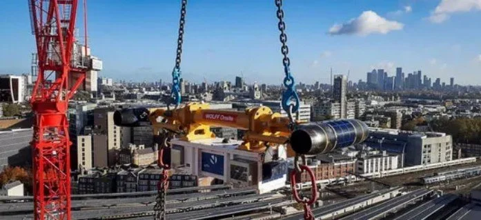 Construction crane lifting a large pipe against city skyline with tall buildings and blue sky.