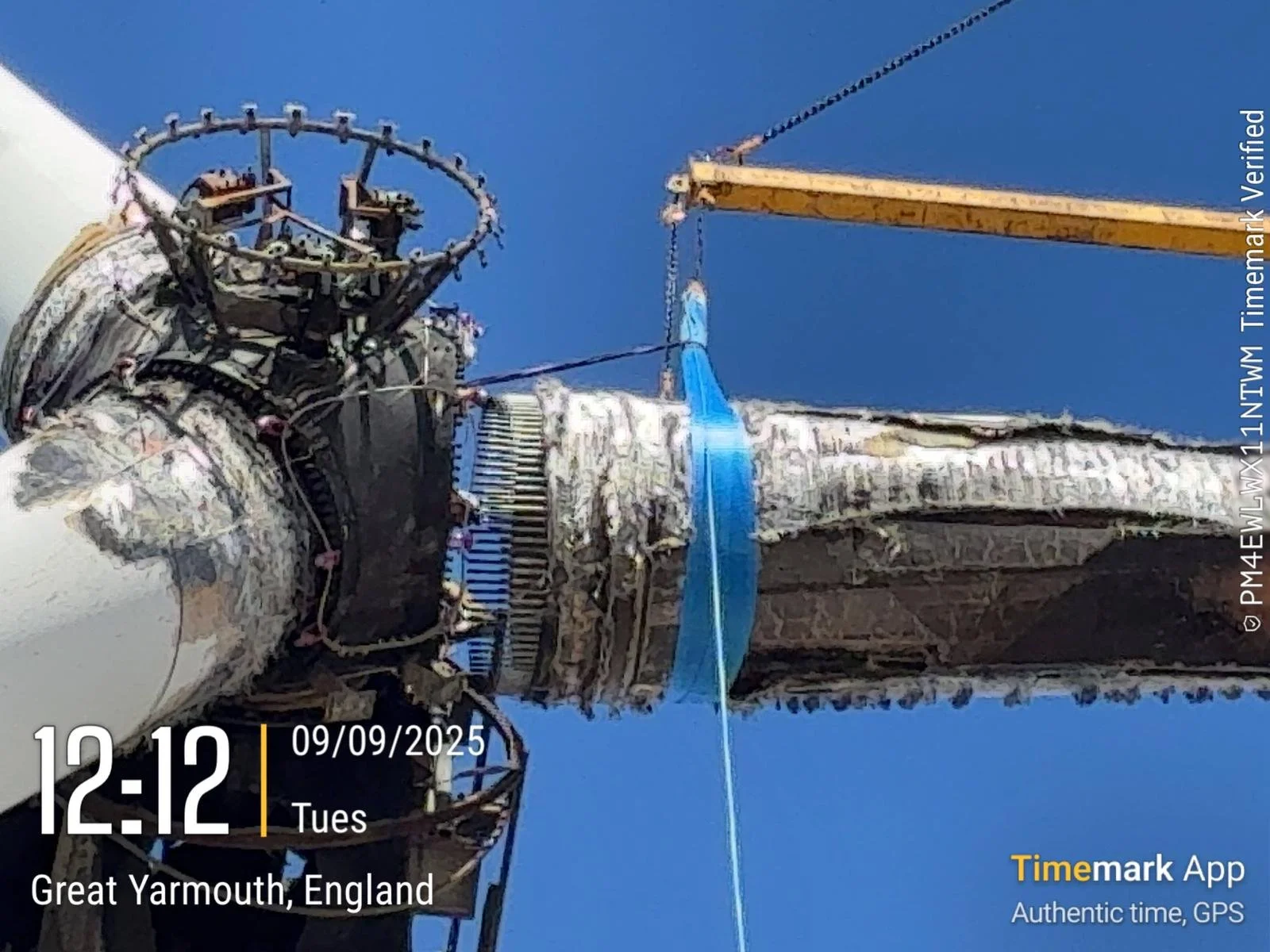 Close-up of a spacecraft's hardware with a yellow crane lifting a blue strap, set against a bright blue sky, with sundry mechanical parts and structures visible.