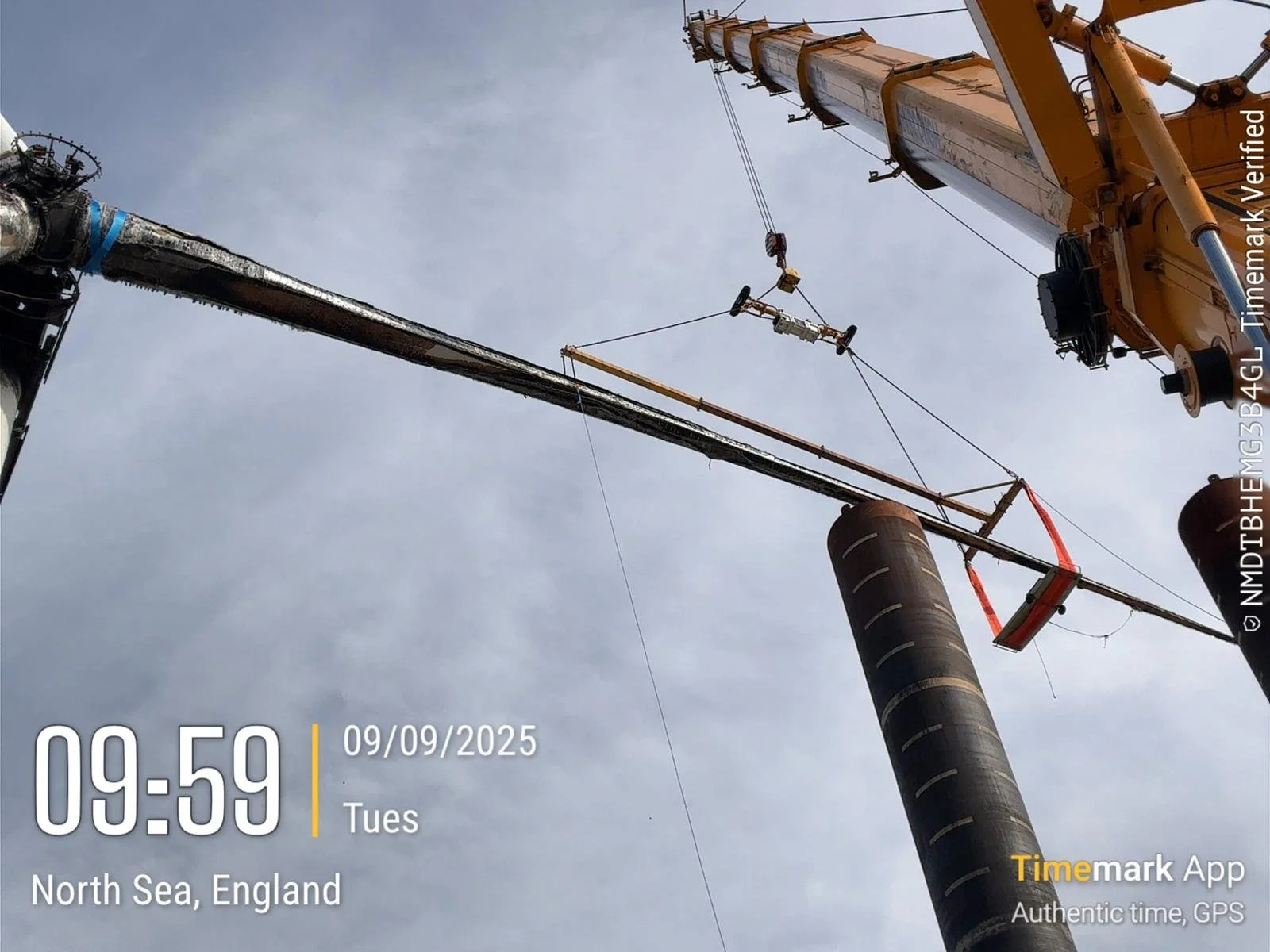 Construction workers are using a crane to lift and install a large utility pole on a cloudy day in North Sea, England. A worker is standing on top of the crane's arm, and the crane is positioned near an existing pole.