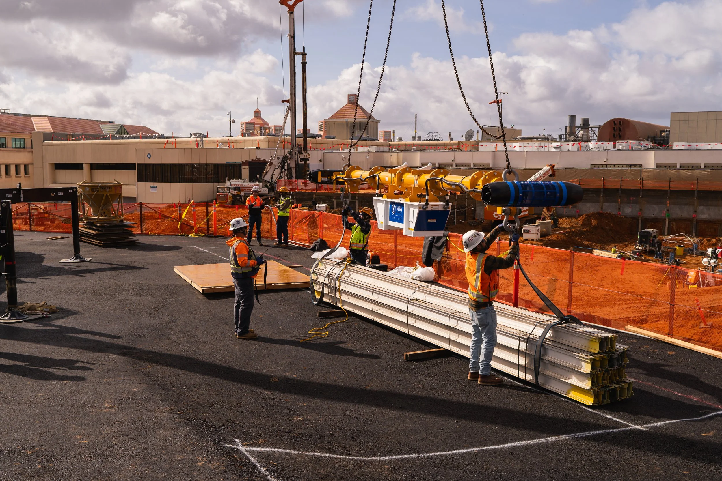 Construction workers in safety gear installing a large piece of machinery on a construction site with orange safety barriers, with a cityscape and partly cloudy sky in the background.