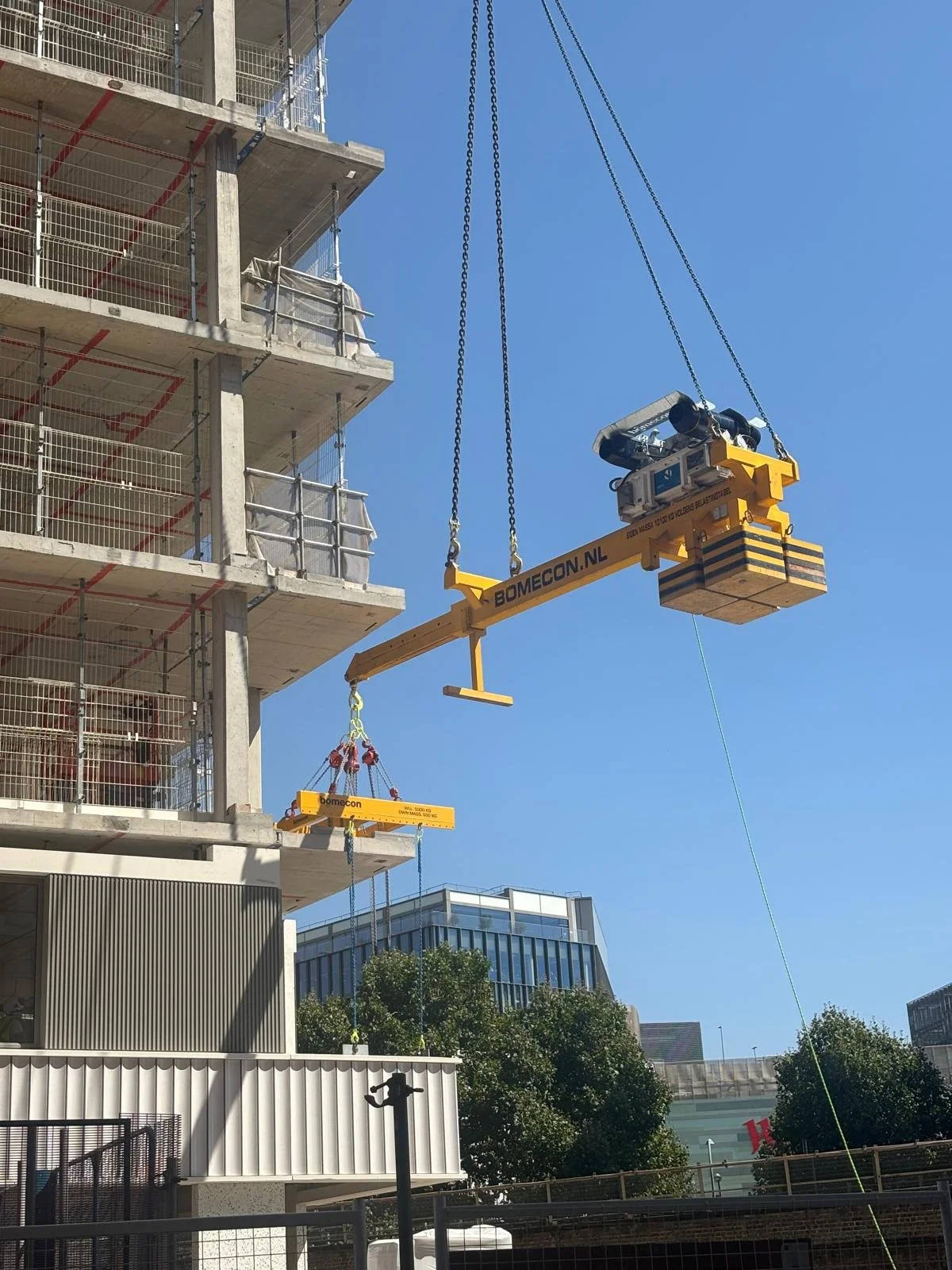 Construction site with a building under construction on the left and a yellow crane lifting a large platform. The building has several floors with open concrete slabs and safety railings. There are some trees and modern buildings in the background un