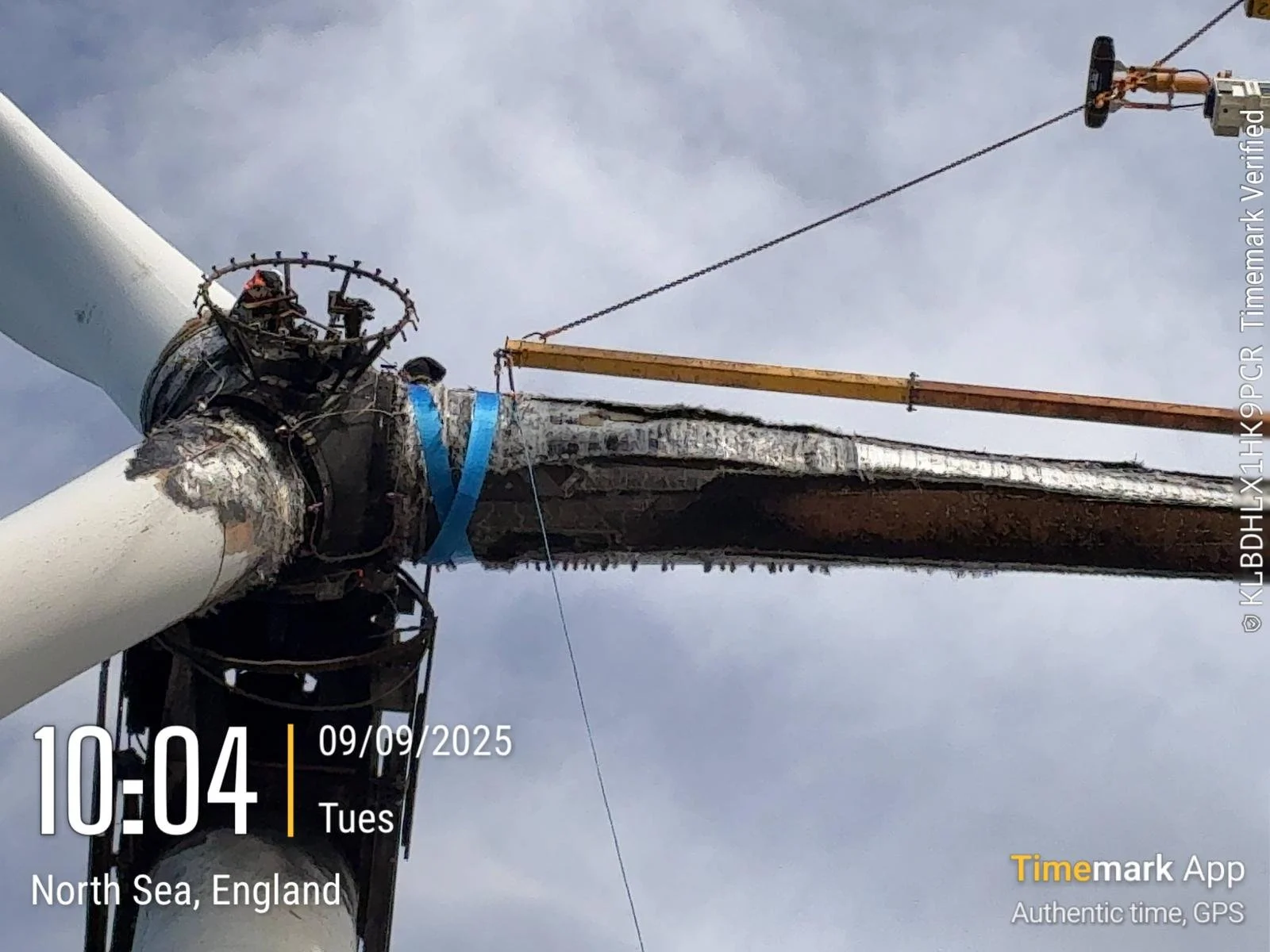 Close-up view of a wind turbine maintenance or construction process showing part of the turbine tower with some blades and the nacelle at the top. A crane is visible, aiding in the work.