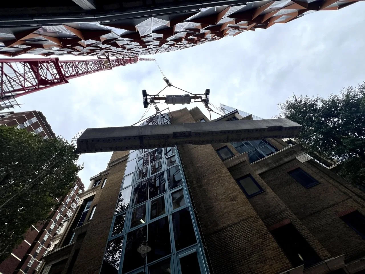 View looking up at construction crane lifting a large building panel, surrounded by tall buildings with brick and glass exteriors, and trees on the side, under a cloudy sky.