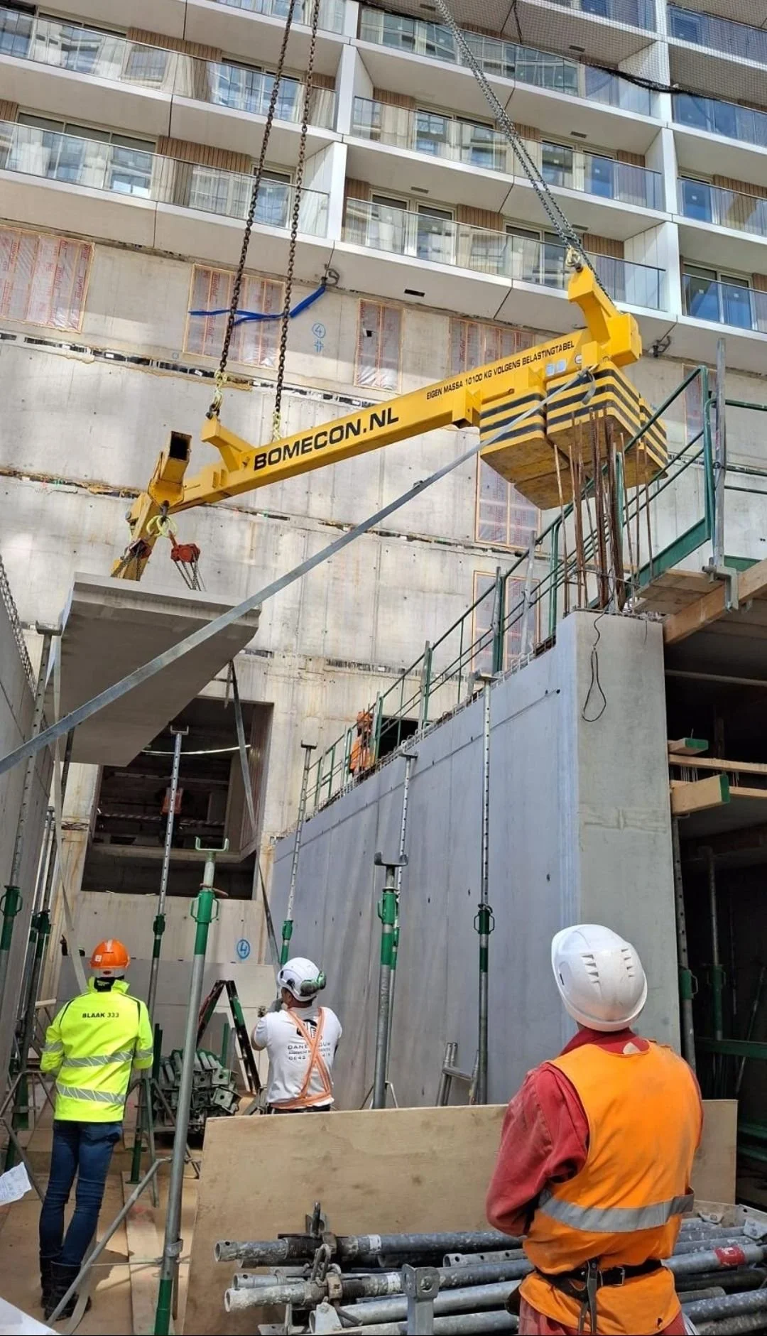 Construction site with workers in safety gear, scaffolding, and a large yellow crane lifting construction materials against a multi-story building.
