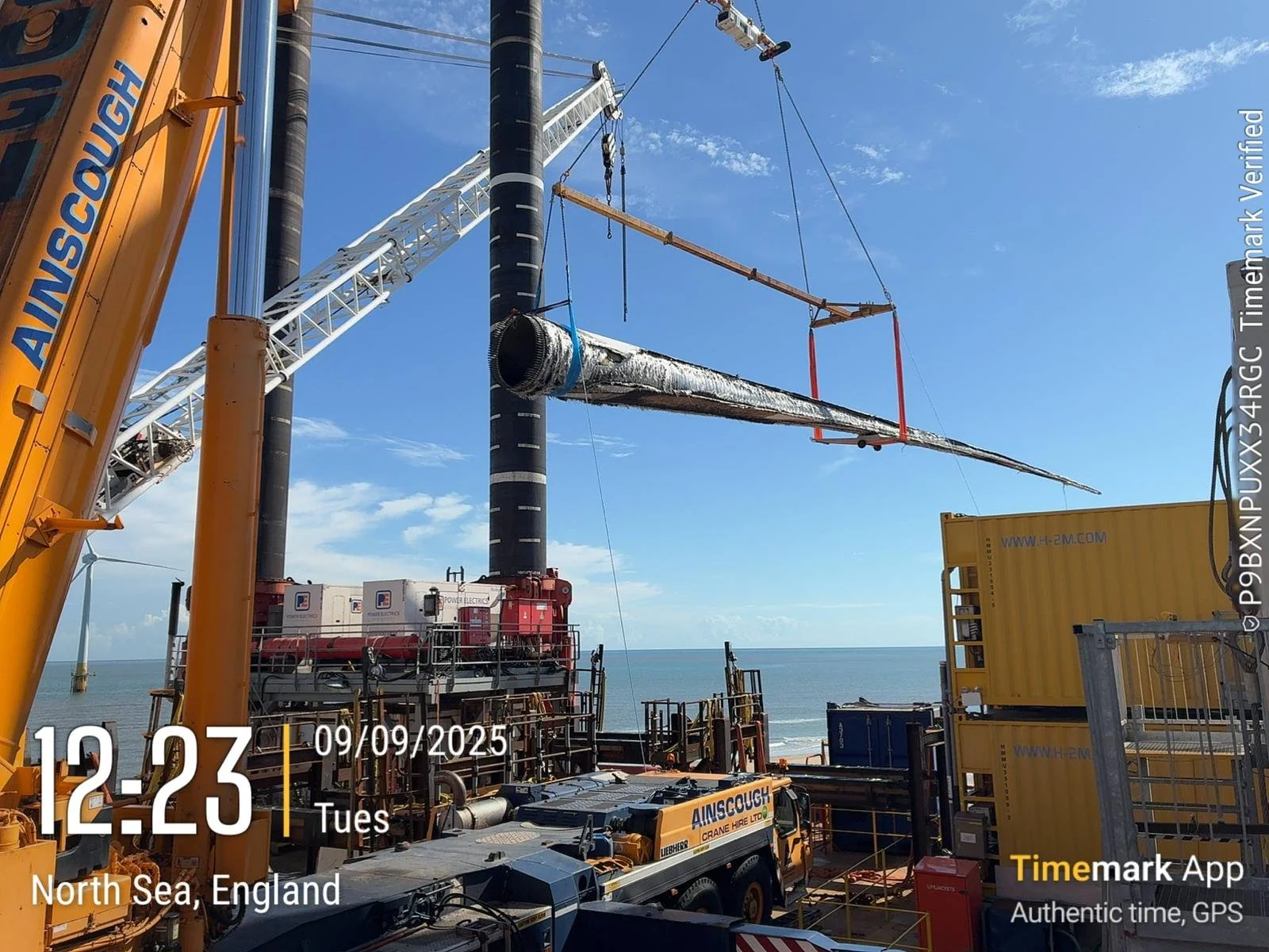Construction site on the North Sea coast in England with large cranes installing a wind turbine blade, blue sky, and ocean in the background. The scene includes machinery and containers, with a timestamp showing 12:23 PM on September 9, 2025.