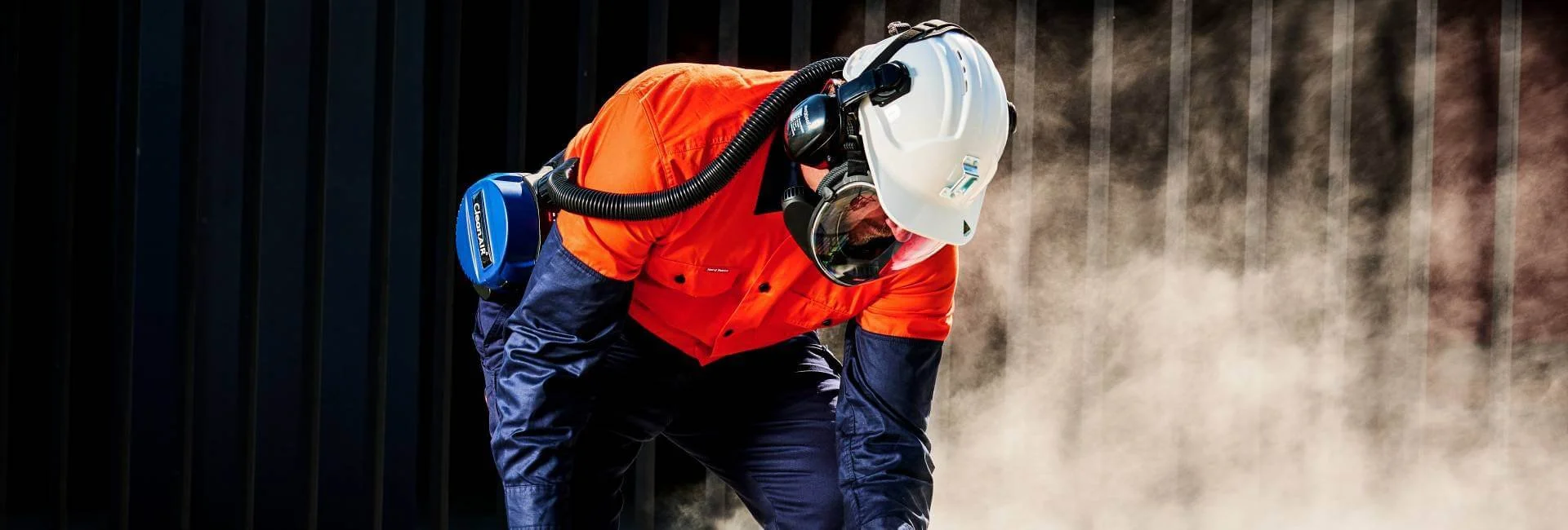 Worker in orange and navy protective gear, wearing a white helmet and respirator, performing a task that produces dust.