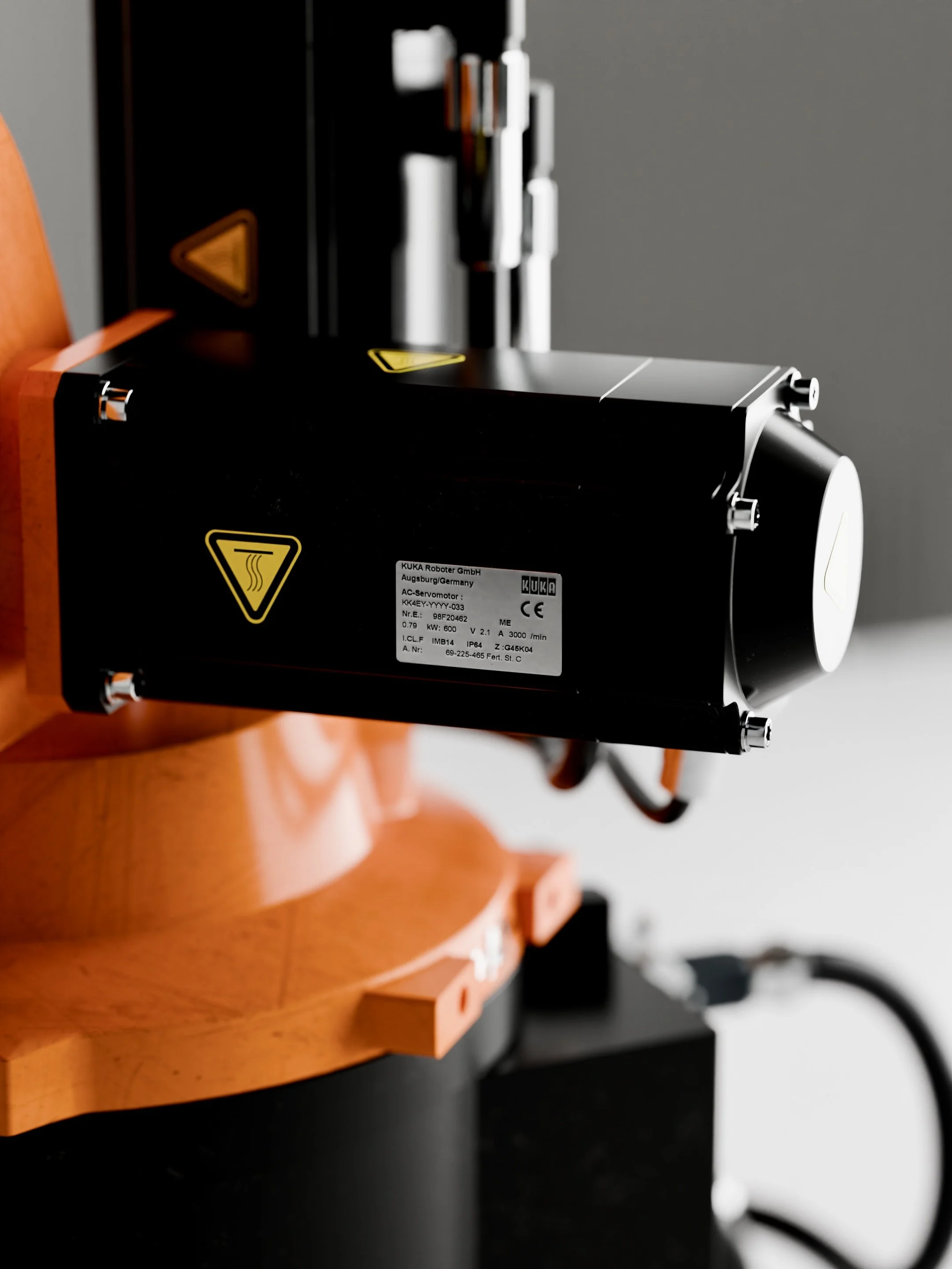 Close-up of a black and orange industrial machine part with a white label containing technical details. The machine appears to be part of a robotic or automation system.