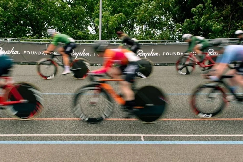 Cyclists racing on a velodrome track, blurred motion, Rapha Foundation signage in background.