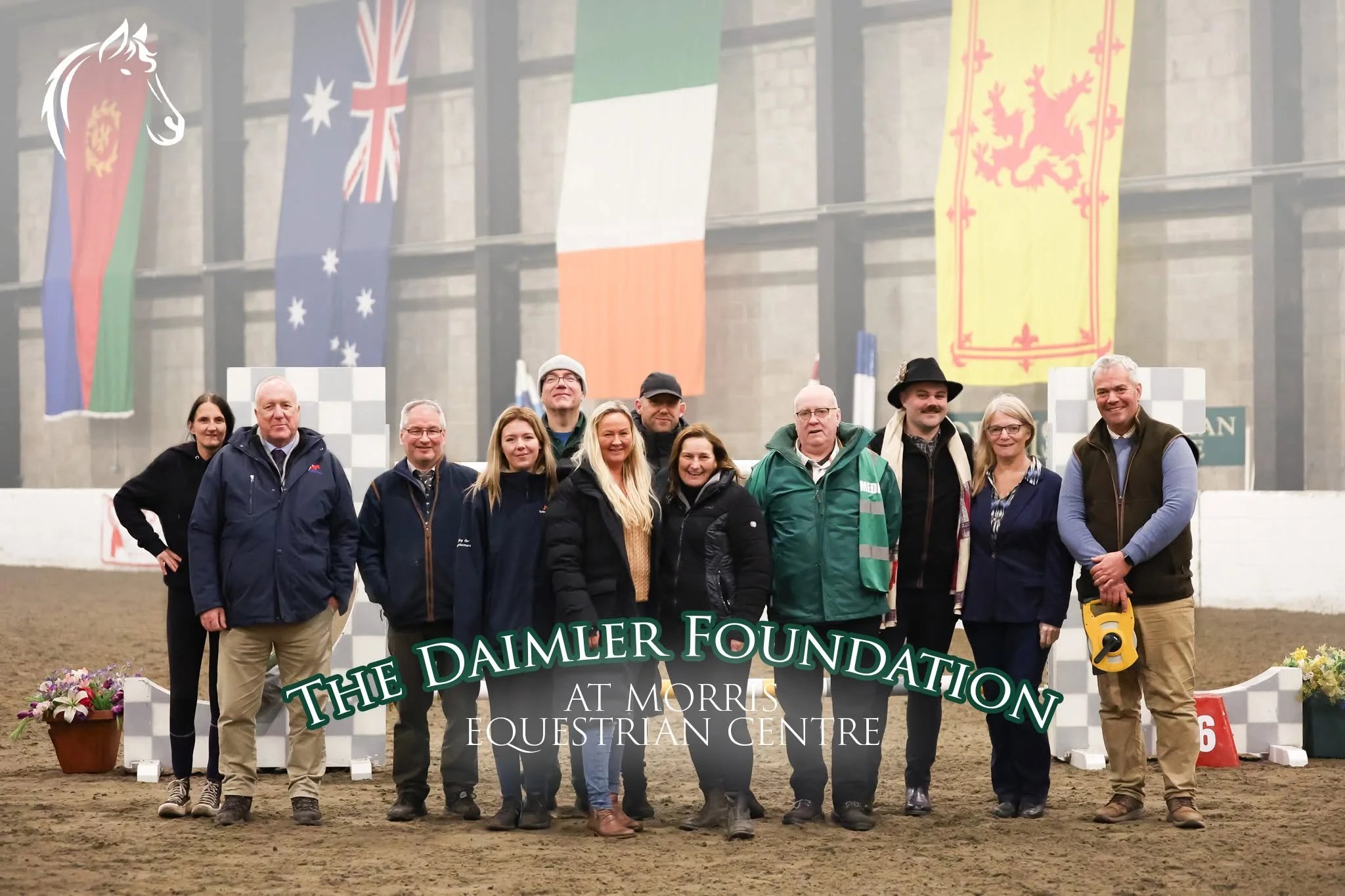 Group of people standing together on an indoor equestrian arena, with flags hanging on the wall behind them, including flags of Australia, Ireland, and other regions. The group is smiling and dressed in casual and equestrian clothing. A sign reading 'The Daimler Foundation at Morris Equestrian Centre' is visible at the bottom of the image.