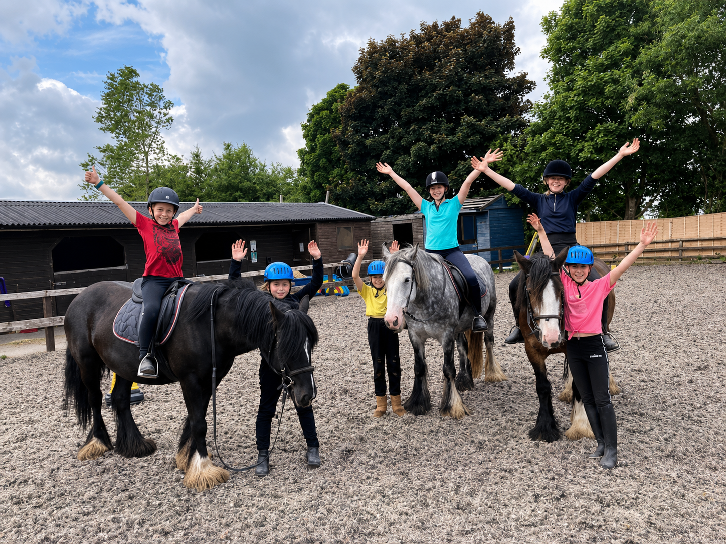 Group of five children riding horses at an outdoor equestrian riding area. Three children are on horseback, while two are standing beside their horses, all wearing helmets and casual clothing. The children are smiling and raising their hands, with a background of trees, buildings, and a cloudy sky.