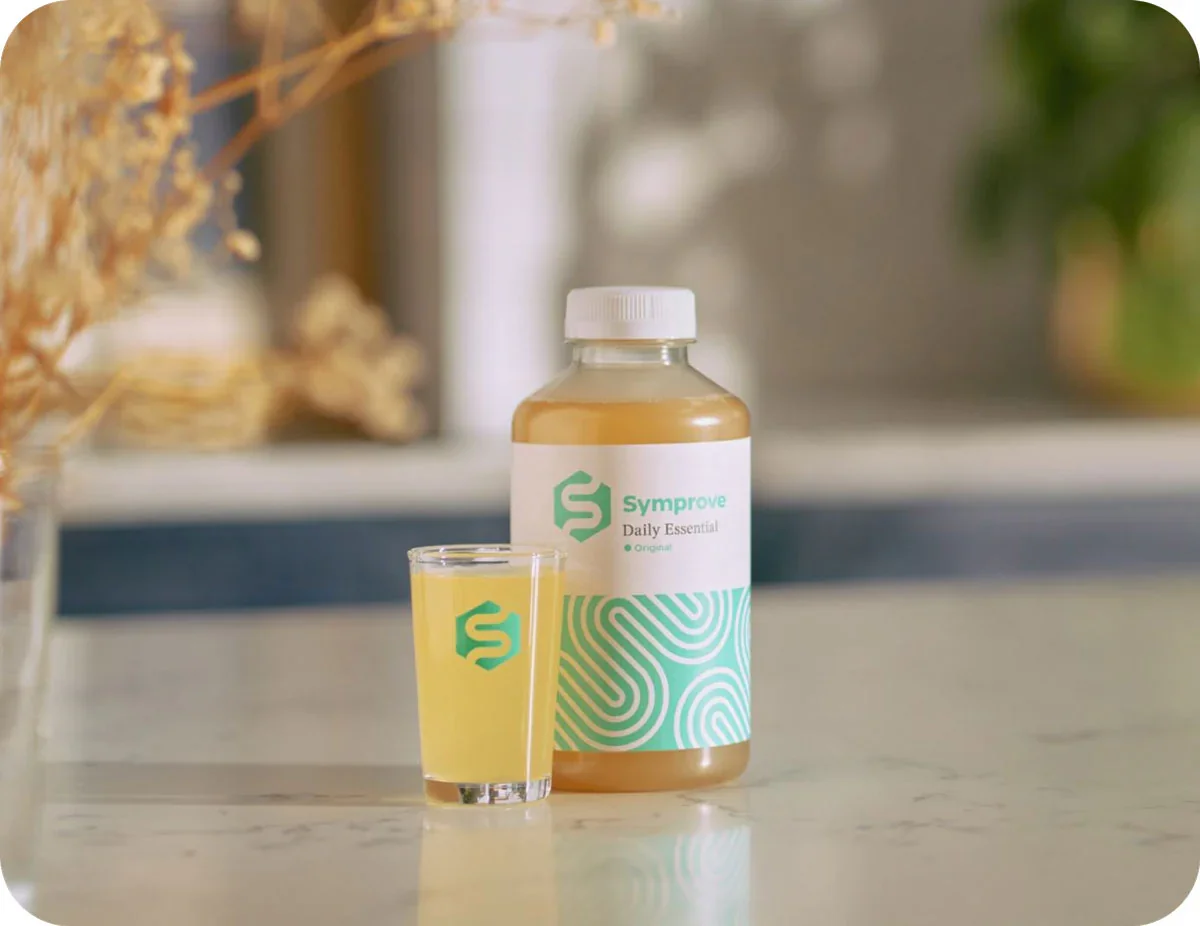 A bottle labeled 'Symprove Daily Essential' standing next to a glass filled with a yellow liquid, both placed on a marble surface in a bright indoor setting.