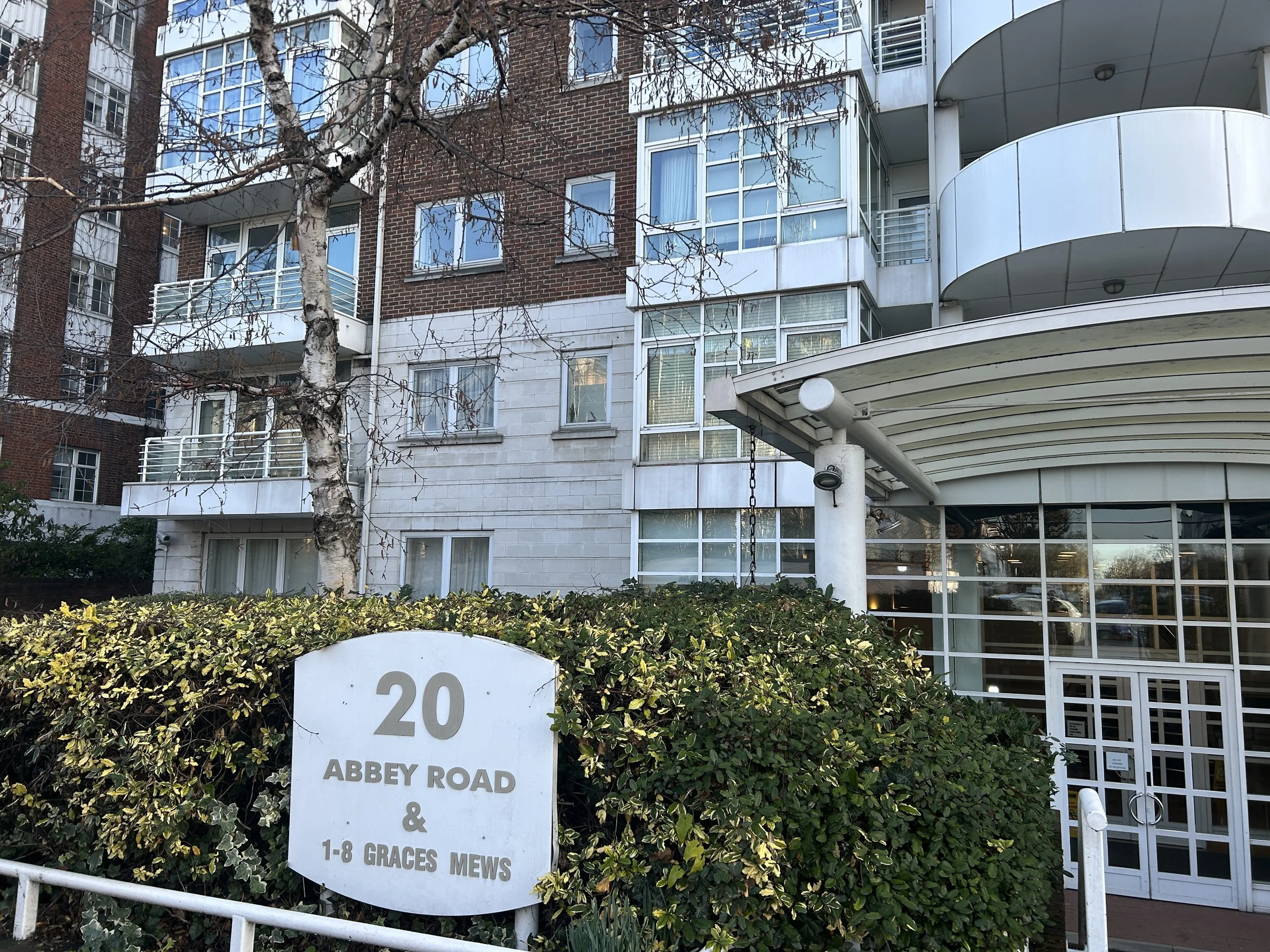 Multi-story residential building with glass balconies and large windows, a tree with bare branches, and a white sign in the front reading '20 Abbey Road & 1-8 Graves Mews'.