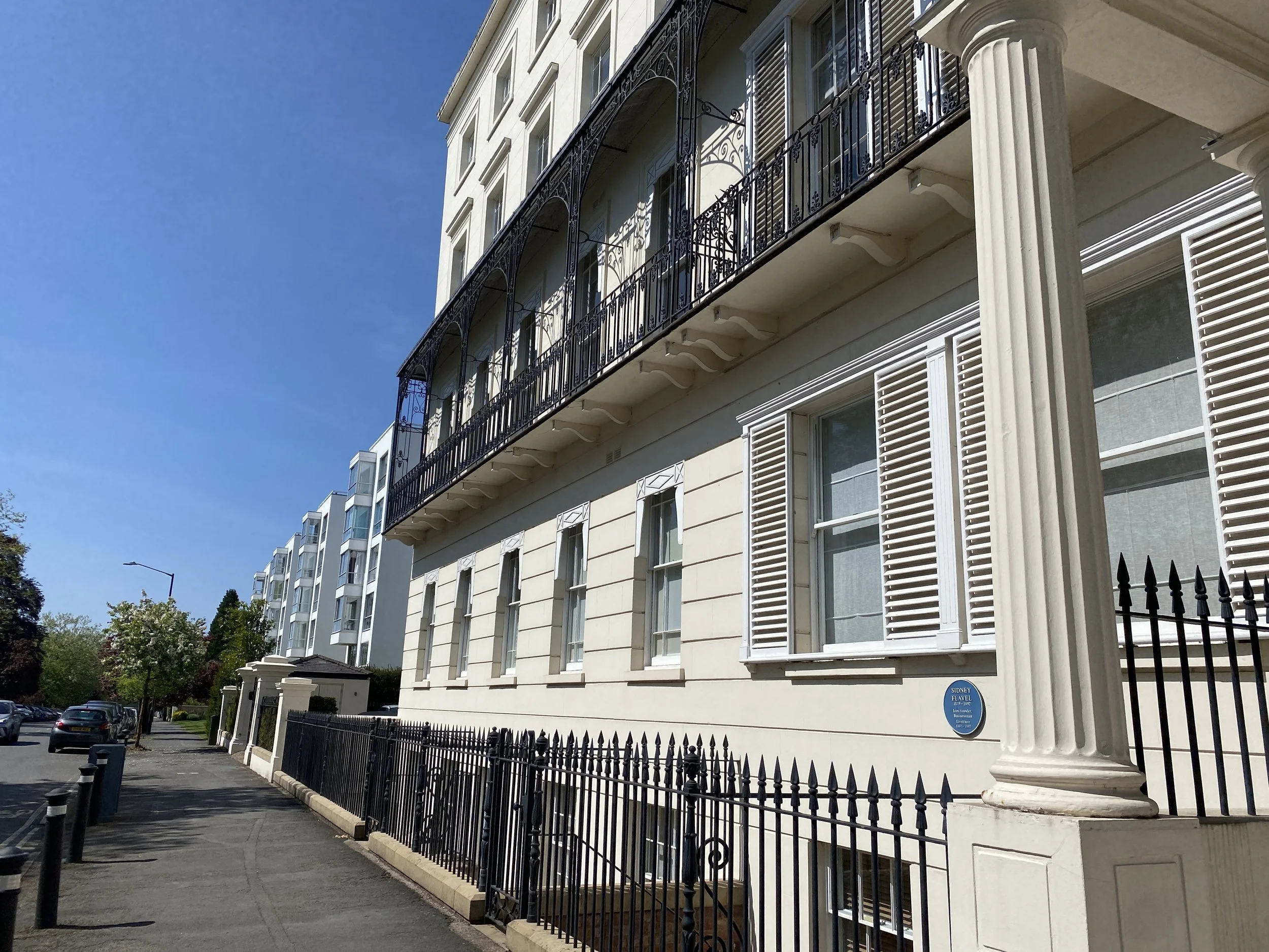A white multi-story building with decorative black wrought iron balconies and window shutters, with a black iron fence along the sidewalk. A street with parked cars and trees is visible in the background under a clear blue sky.
