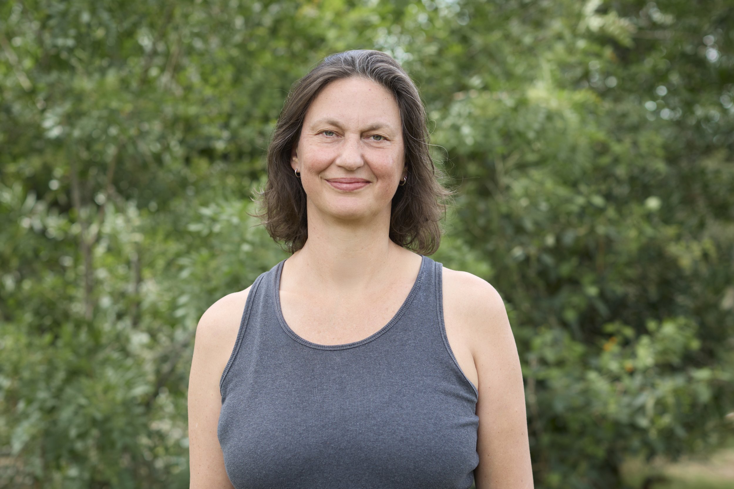 Mujer joven con cabello castaño, camiseta gris y pendientes pequeños, sonriendo en un entorno natural con árboles verdes de fondo.