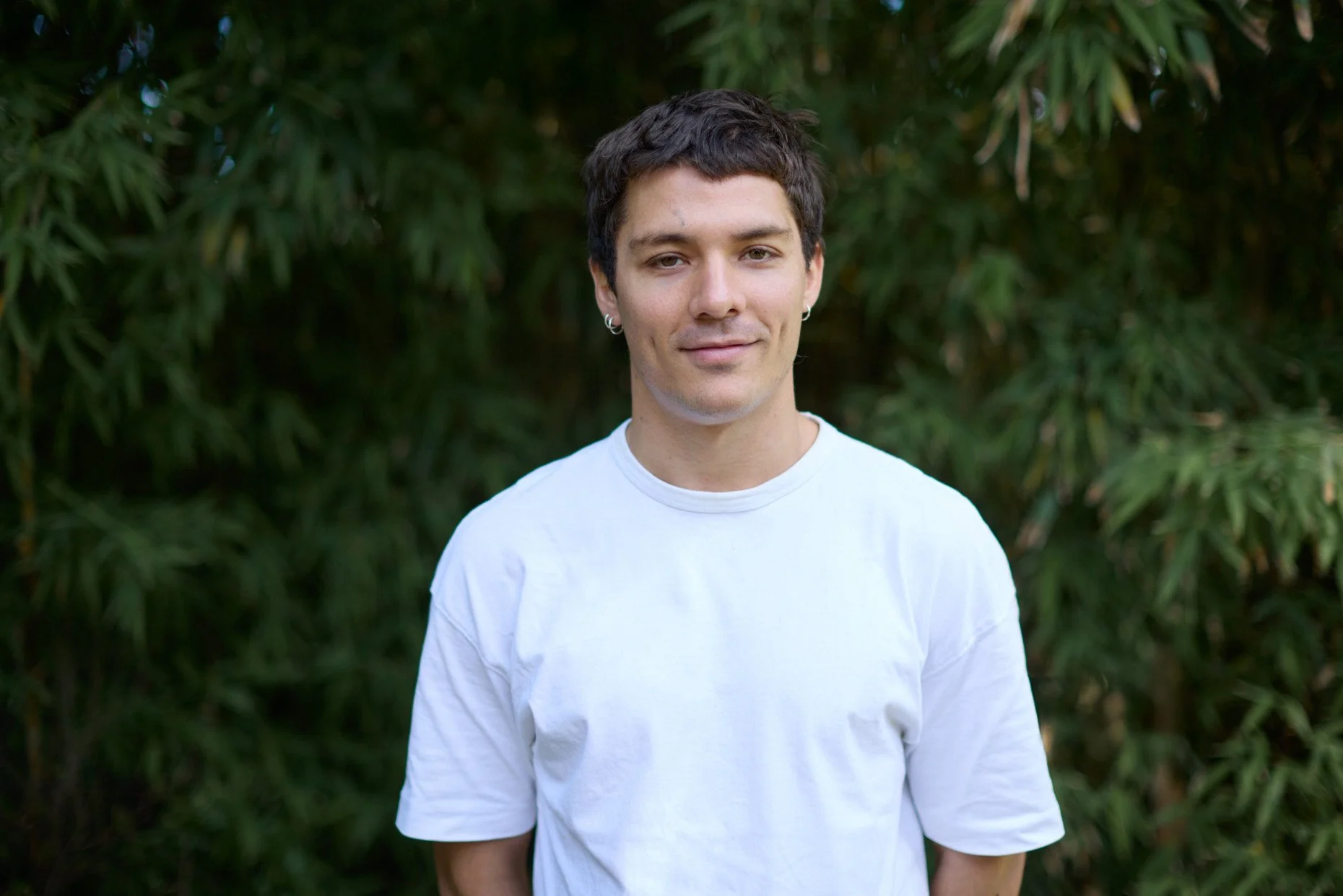 Retrato de un hombre joven con camiseta blanca frente a un fondo de vegetación verde.