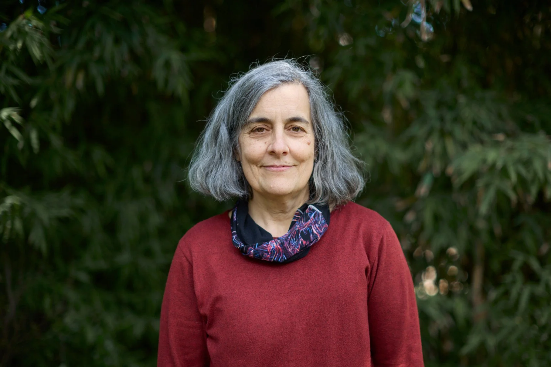 Una mujer mayor con cabello gris y ondulado, vestida con un suéter rojo y un pañuelo con patrones en el cuello, posando frente a un fondo de vegetación verde.