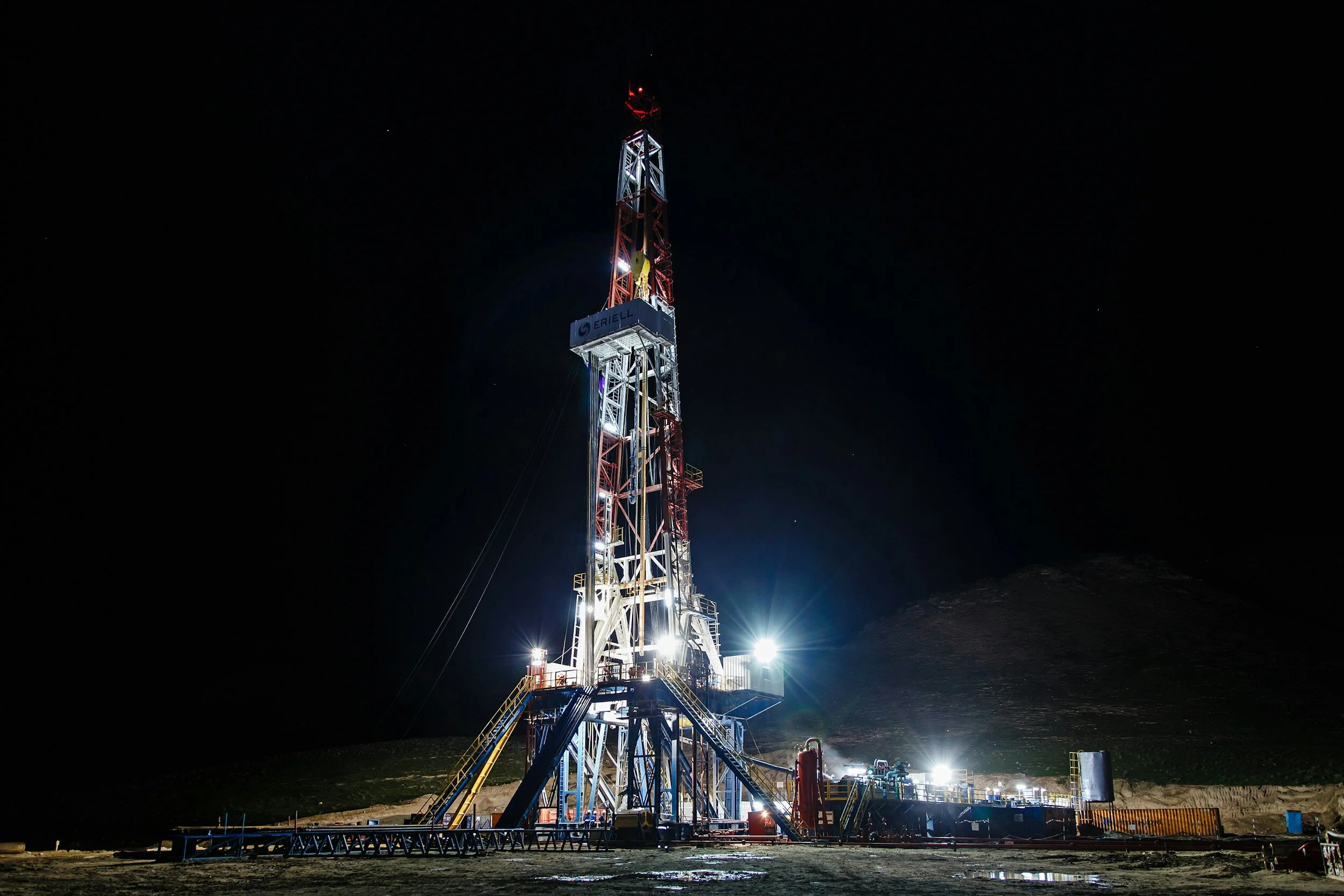 Illuminated oil drilling rig at night with lights, set against a dark sky.