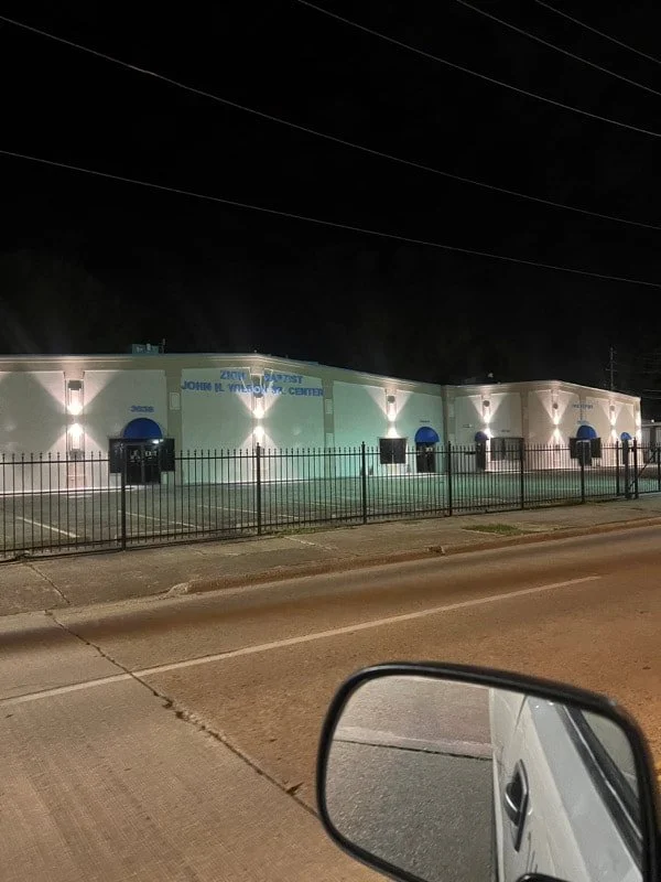A large building with blue accents and a sign that reads "John H. Winsett Center." It's night, and the building is illuminated. There's a metal fence in front and a vehicle's side mirror visible in the foreground.