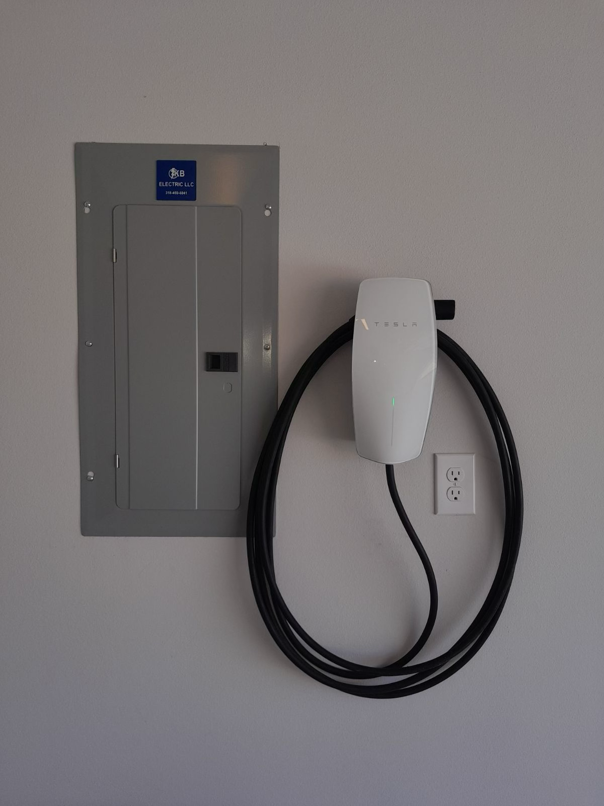 Tesla wall charger with coiled cable next to an electrical panel on a wall.