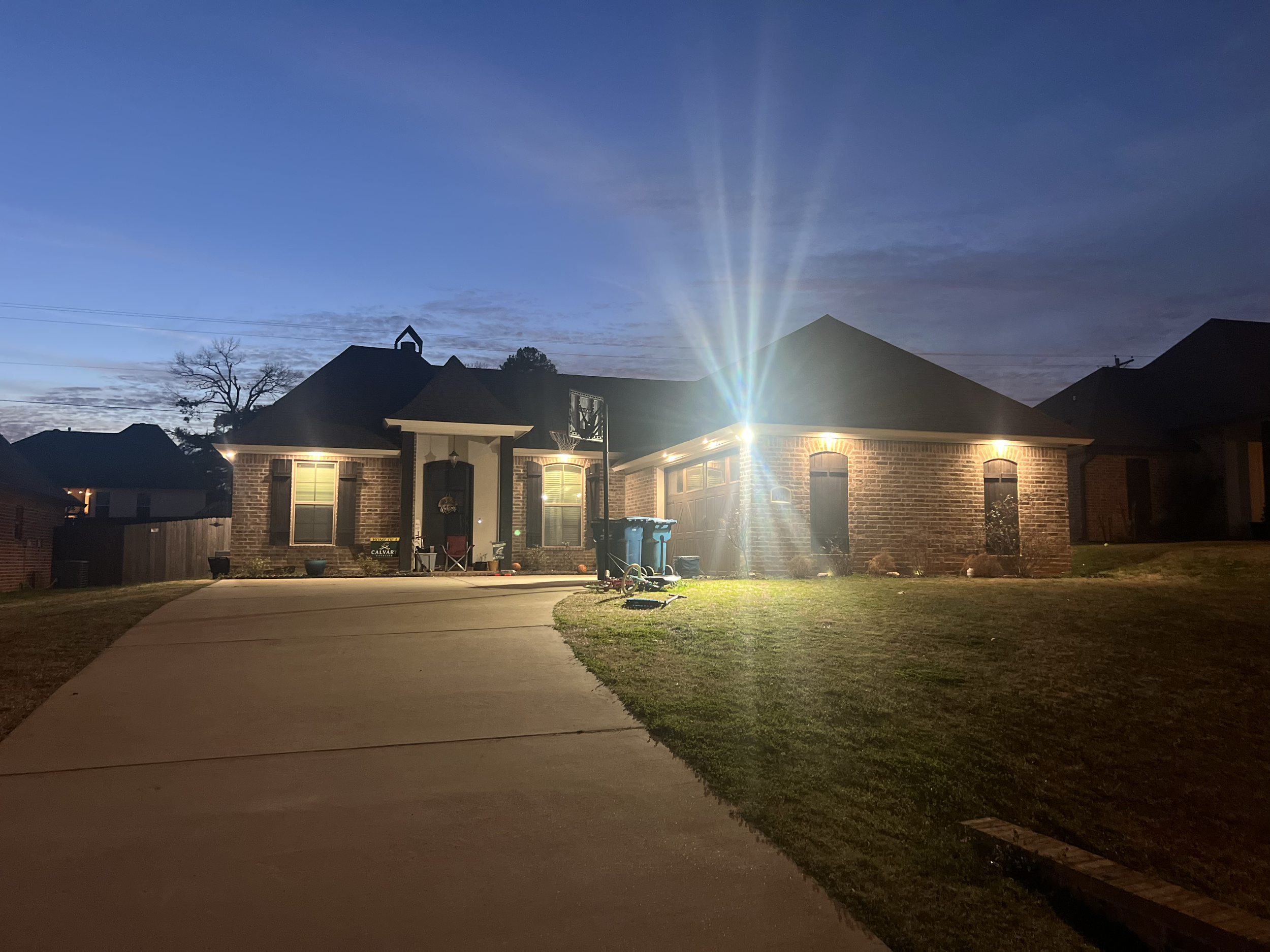 Brick house with a lit driveway at dusk, featuring illuminated outdoor lights, a grassy yard, and a darkening evening sky.