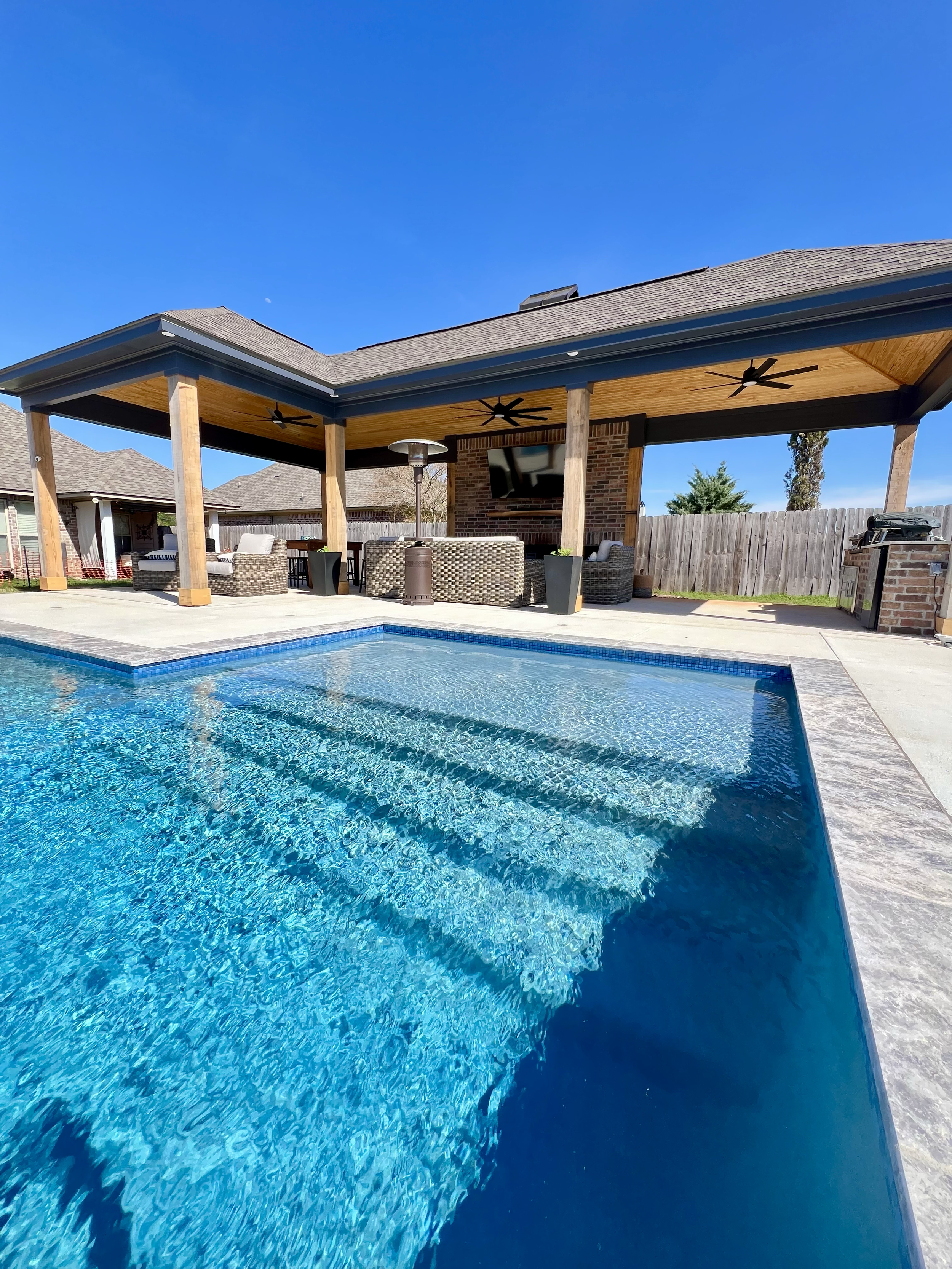 Backyard pool area with a covered patio, outdoor seating, ceiling fans, and a TV under a blue sky.