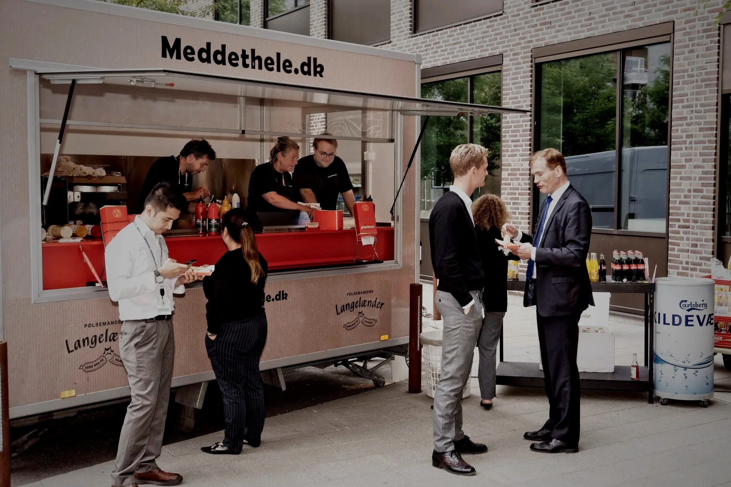 En madvogn med branding, hvor folk bestiller og spiser mad. Der er også nogle personer, der står og taler, mens de spiser. En person i sort outfit arbejder bag disken, og der er en gruppe personer, der står foran og nyder deres mad udendørs.