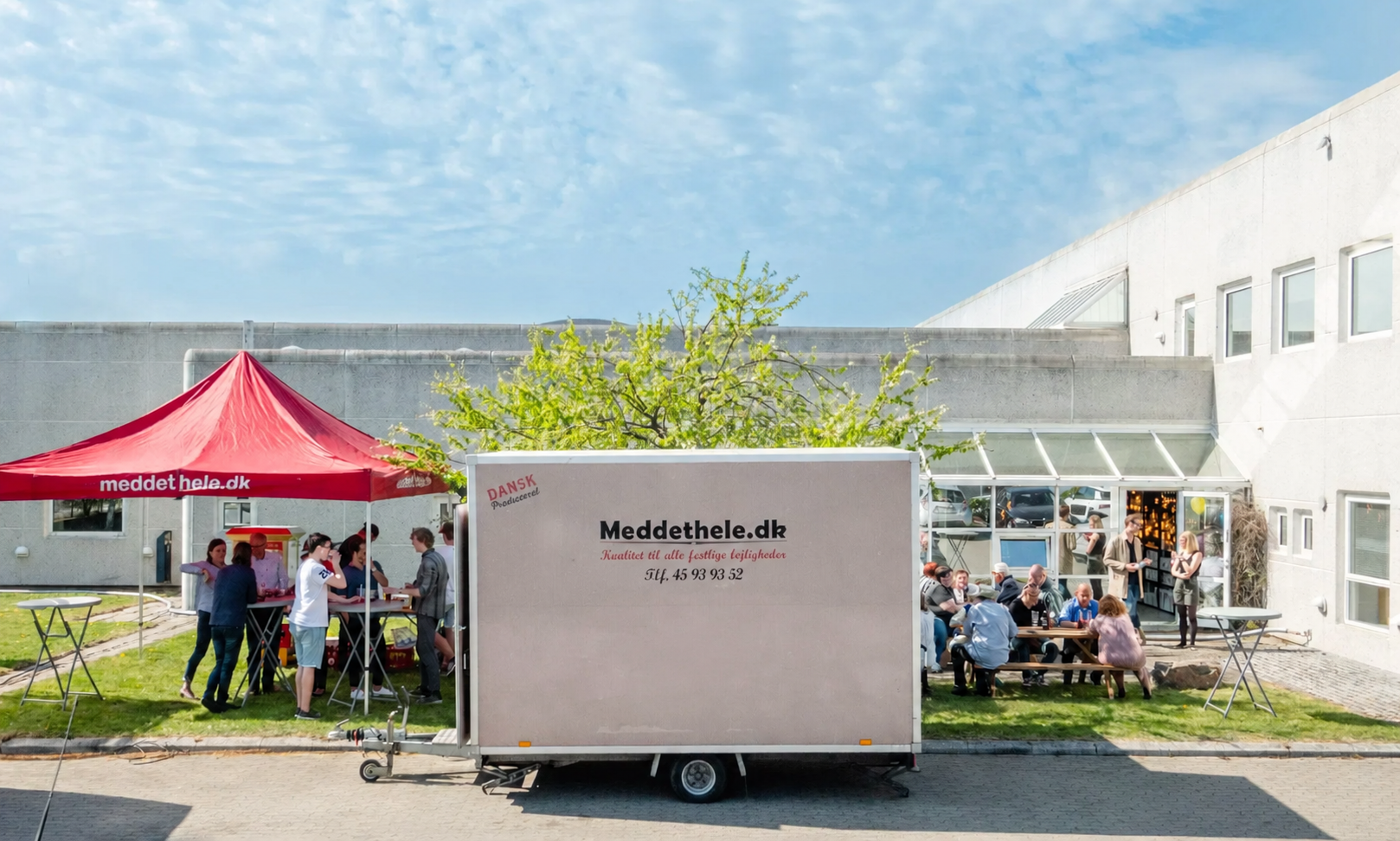 Udendørs madmarked med gruppe mennesker, der sidder og snakker ved borde, og en foodtruck »Meddethle.dk«. Der er en rød telt med teksten »meddet hele.dk«, grønne træer og en bygning med store vinduer i baggrunden.