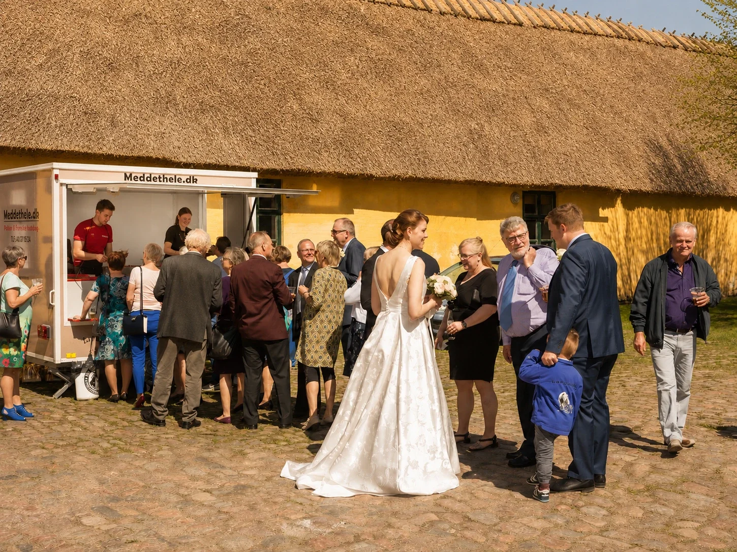 En gruppe mennesker ved en udendørs bryllupsfest, inklusive en brud i hvid kjole, der holder en bouquet, sammen med gæster, der snakker og nyder. Der er en madvogn i baggrunden, og de står på en brostensbelagt plads foran en gul væg under en strålend