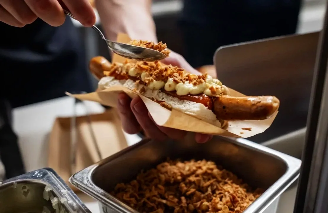 En person holder en hotdog, serveret med mayonnaise, løg og ketchup, og har en pølser i en bolle, mens der er en beholder med ristede løg under.