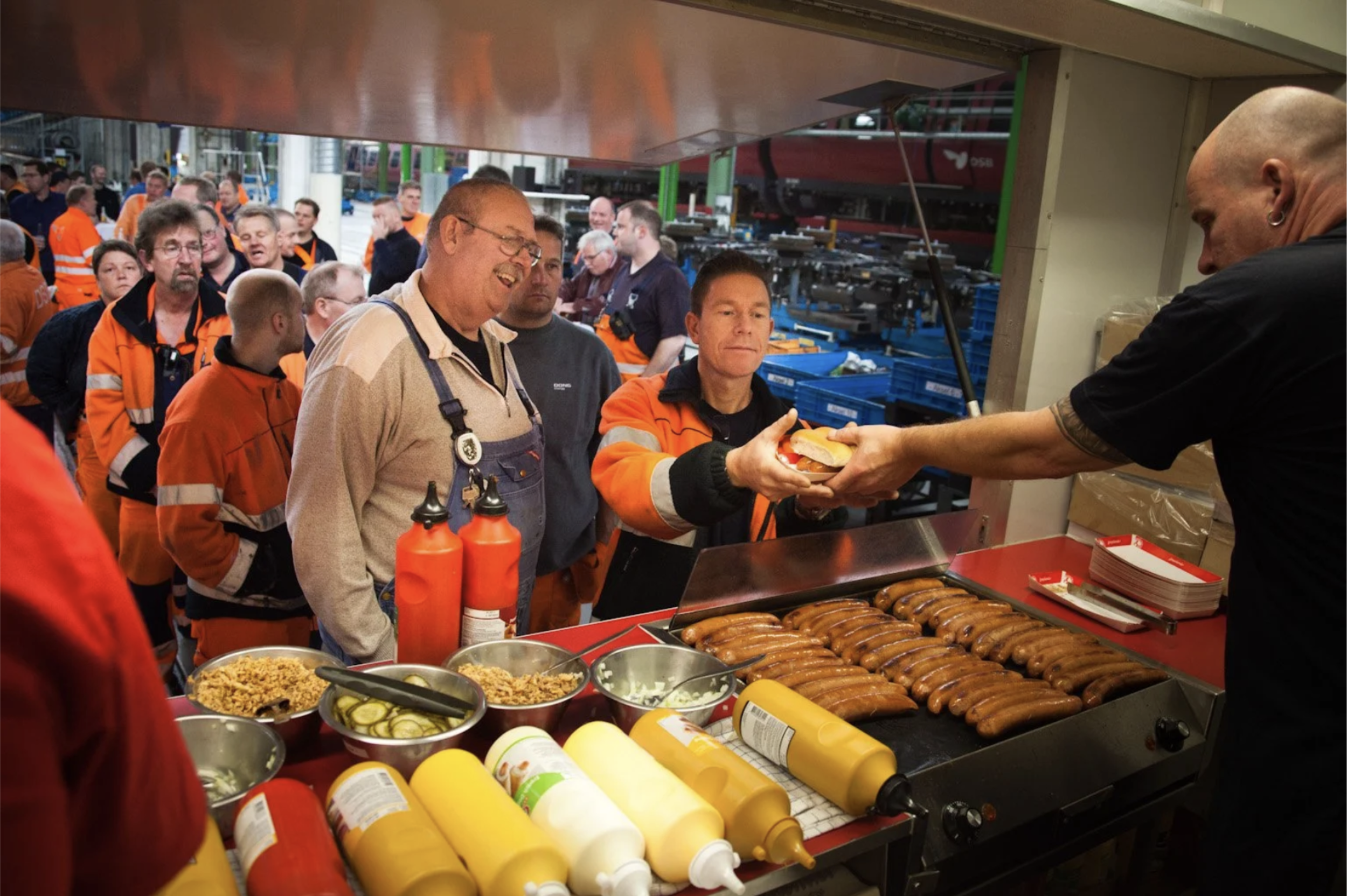 En gruppe mennesker, mange iført orange arbejdstøj, står i kø for at modtage hotdogs hos en madbod, mens nogle andre er i baggrunden.