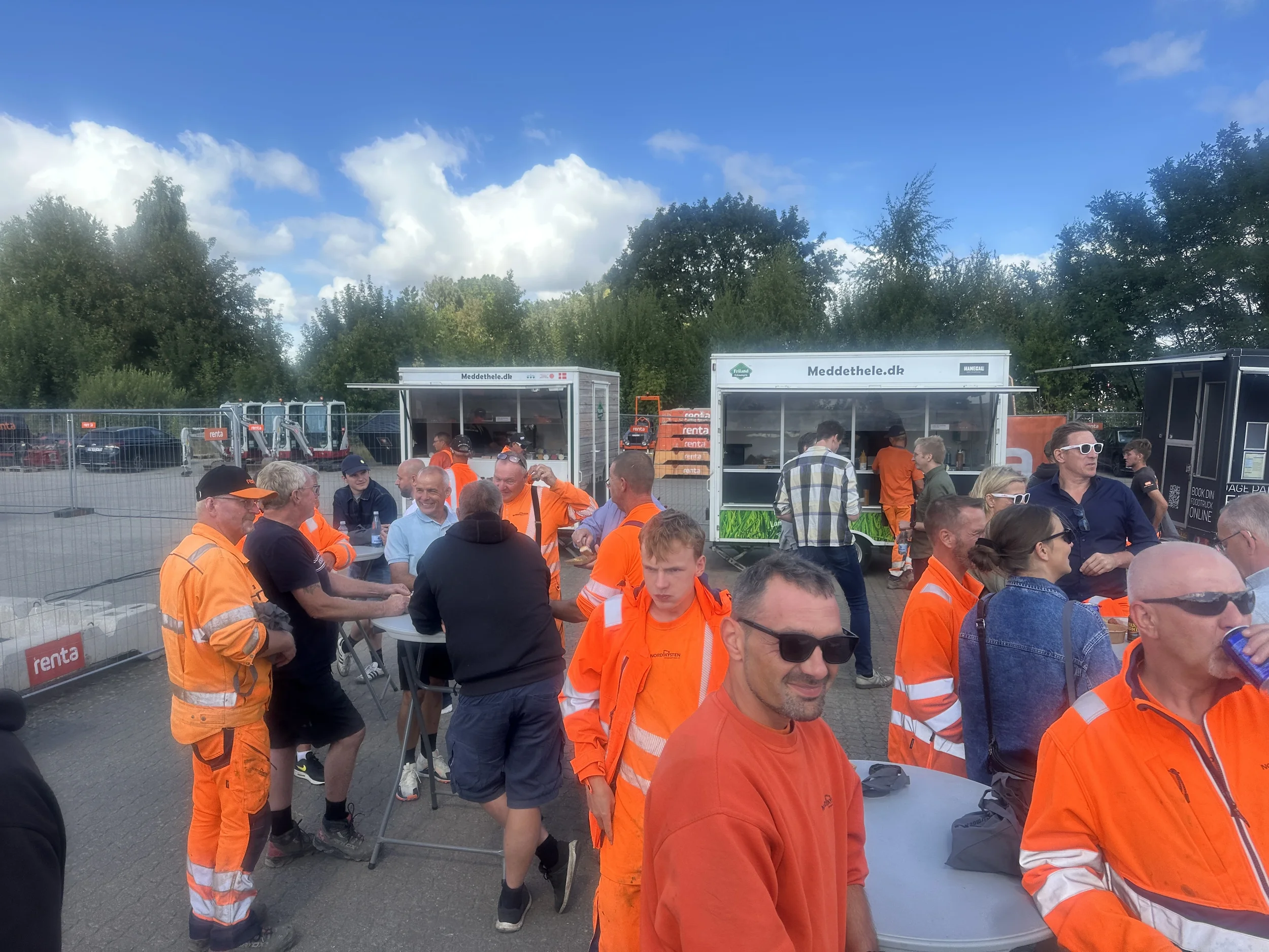 En gruppe mennesker, mange i orange tøj, står og sidder ved borde udenfor, mens de nyder et arrangement. Der er to kørende food trucks i baggrunden, og nogle mennesker venter i kø. Det er en solrig dag med blå himmel og spredte skyer.