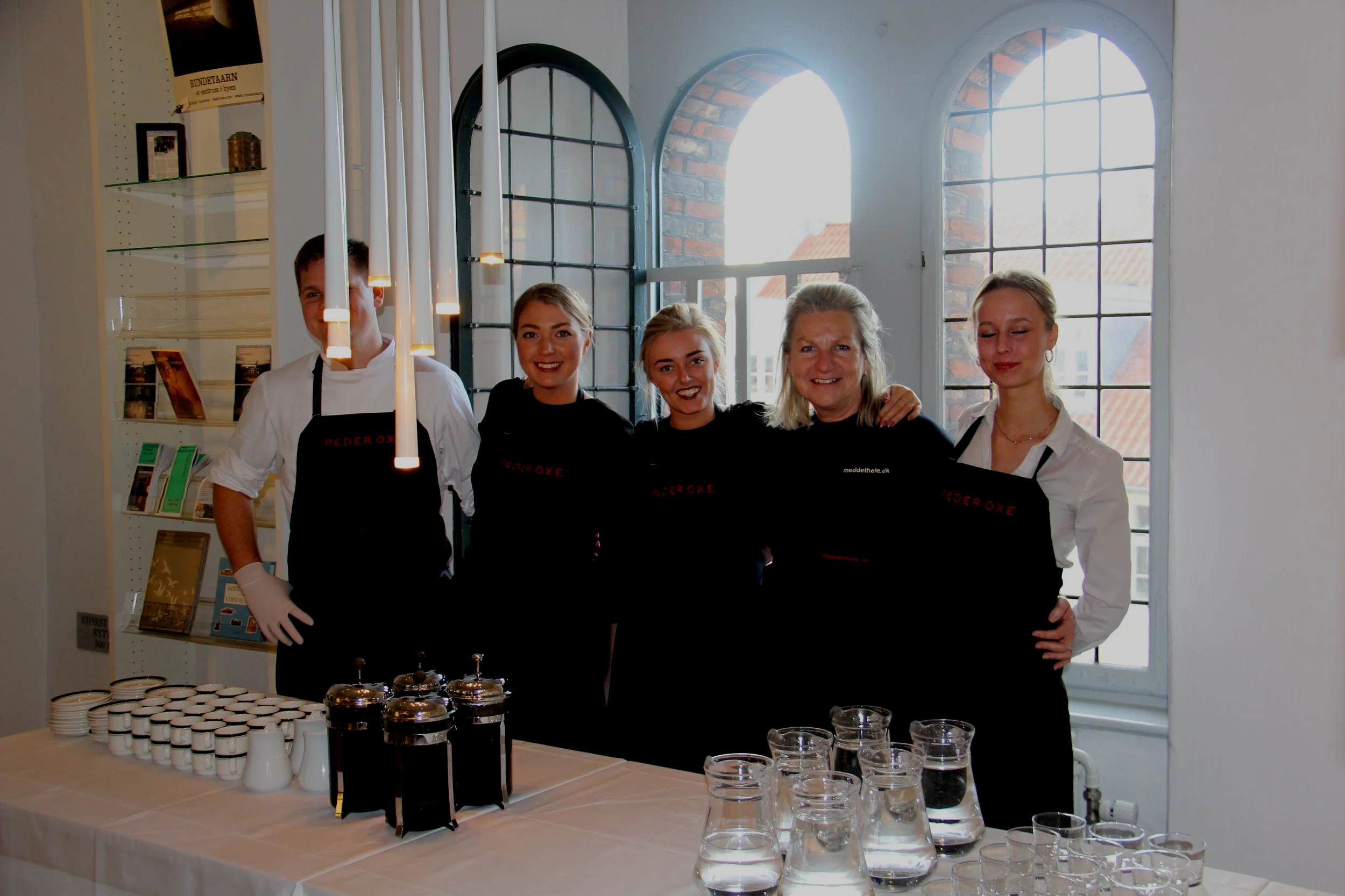 Fem personer står foran et bord med coffee carafes, kopper, glas og vandkander i et lyst rum med store vinduer.