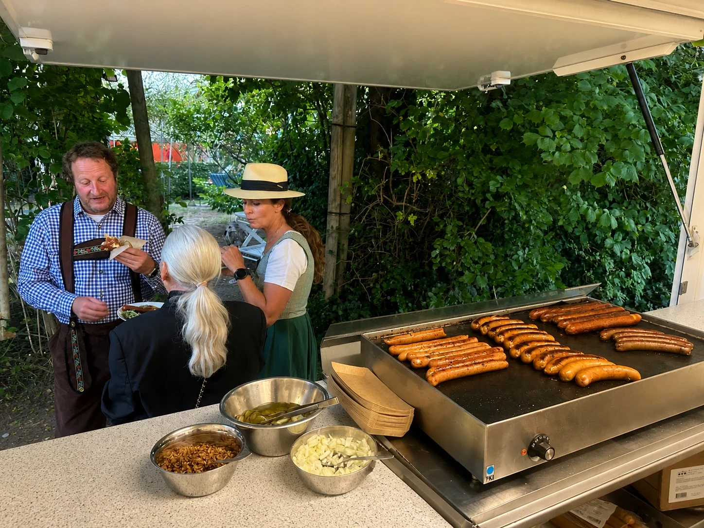 Tre personer, to kvinder og en mand, står ved en grill med pølser, mens de spiser og snakker. Der er tre metalboller med tilbehør som løg, pickles, og pasta-salat. Baggrunden er omgivet af grønne buske.