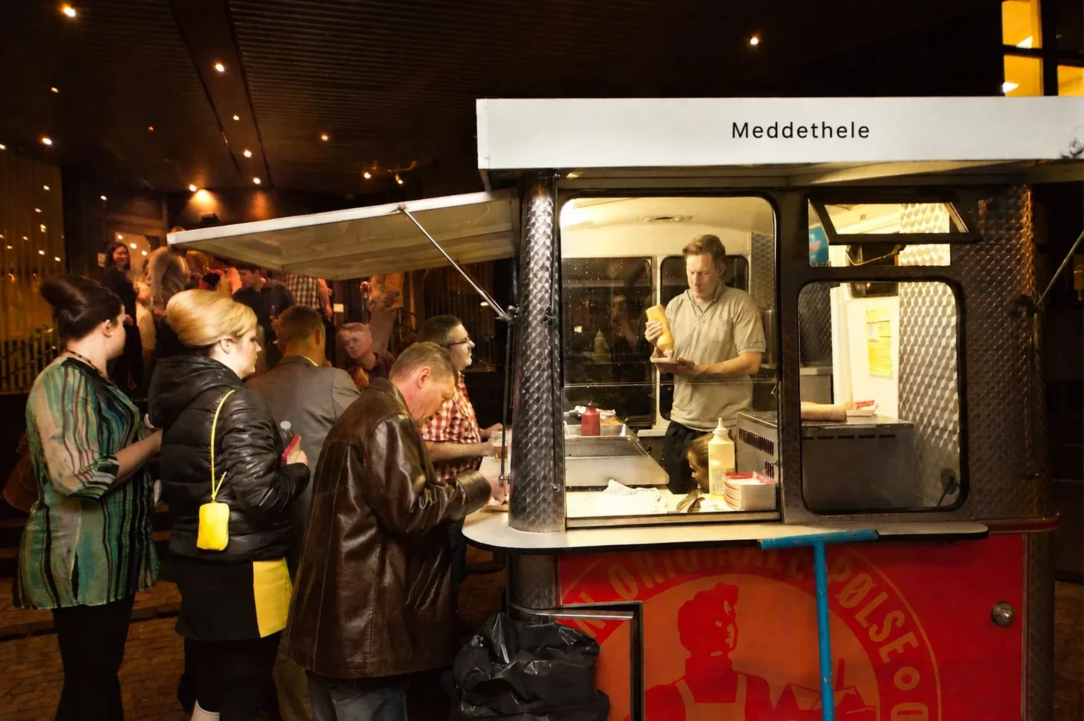 Folk står i kø foran en madvogn, hvor en mand udretter mad. Madvognen er mærket 'Meddheole' og har en sort og rød farve med en logo med en løve.