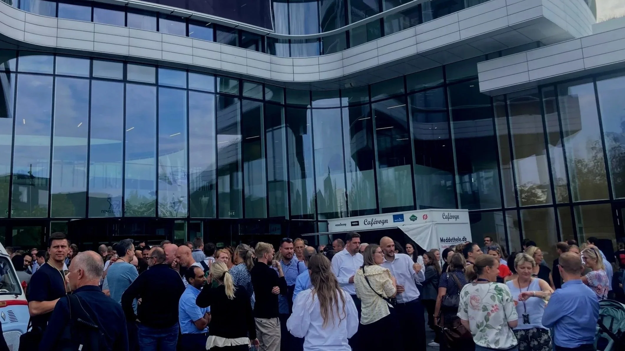 En gruppe mennesker står udenfor en moderne glasbygning, nogle taler, andre venter eller spiser is. Det er en folkesamling foran indgangen.